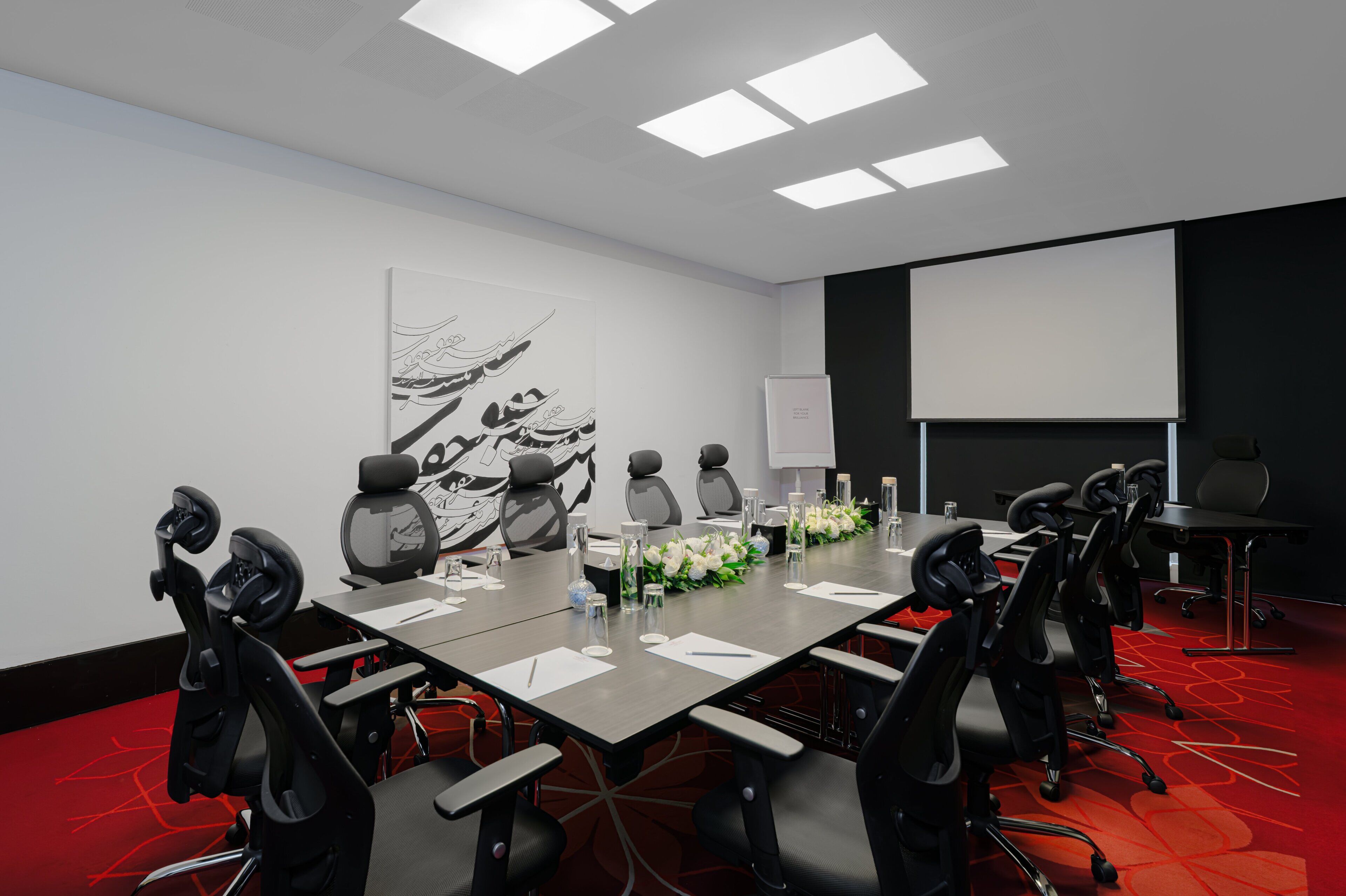 A conference room with a long table, multiple chairs, a projector screen, and a whiteboard.