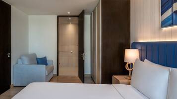 A hotel room with a large bed, a sofa, a bedside table with a lamp, and a door leading to a bathroom.