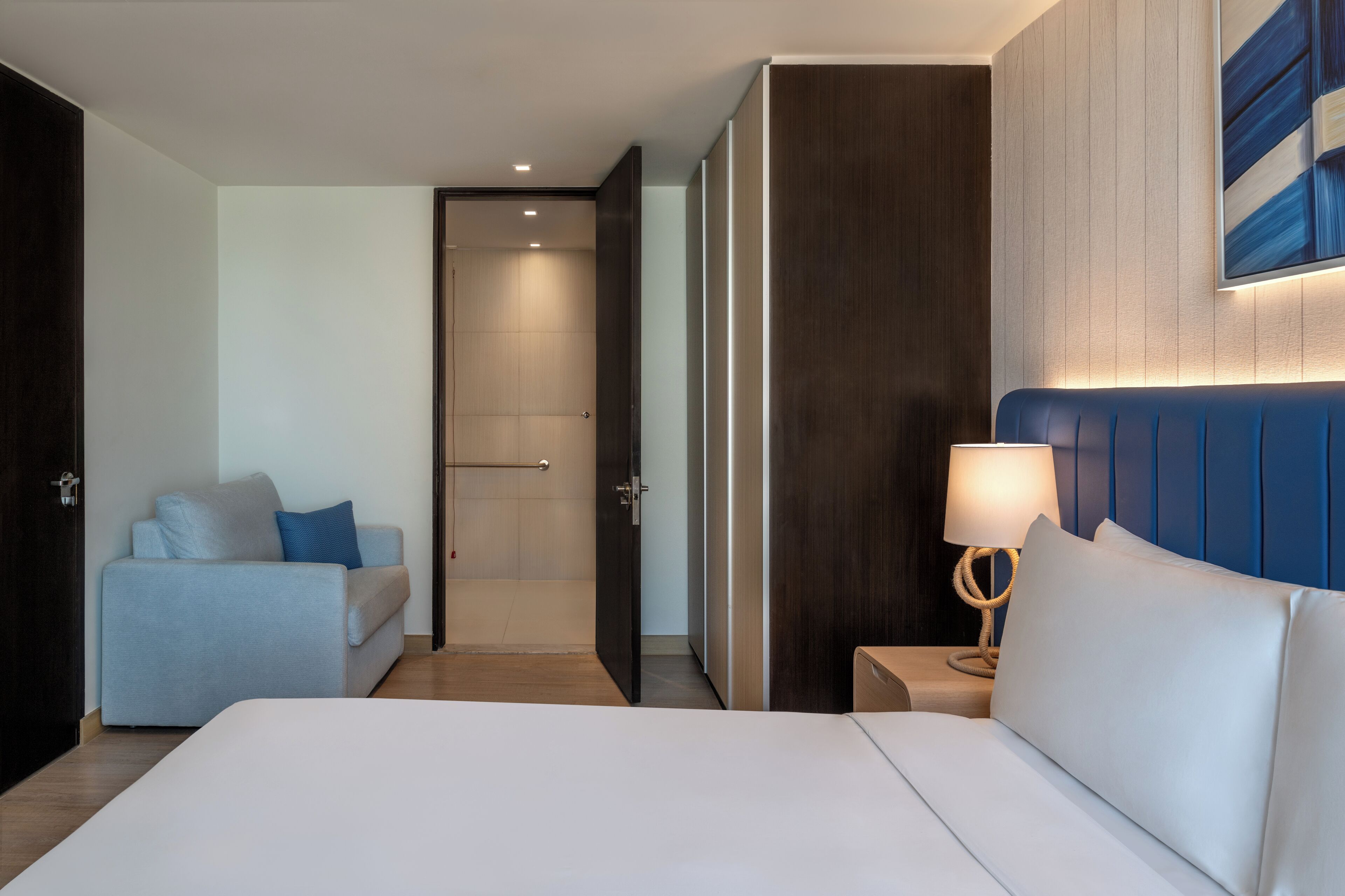 A hotel room with a large bed, a sofa, a nightstand with a lamp, and a door leading to a bathroom.