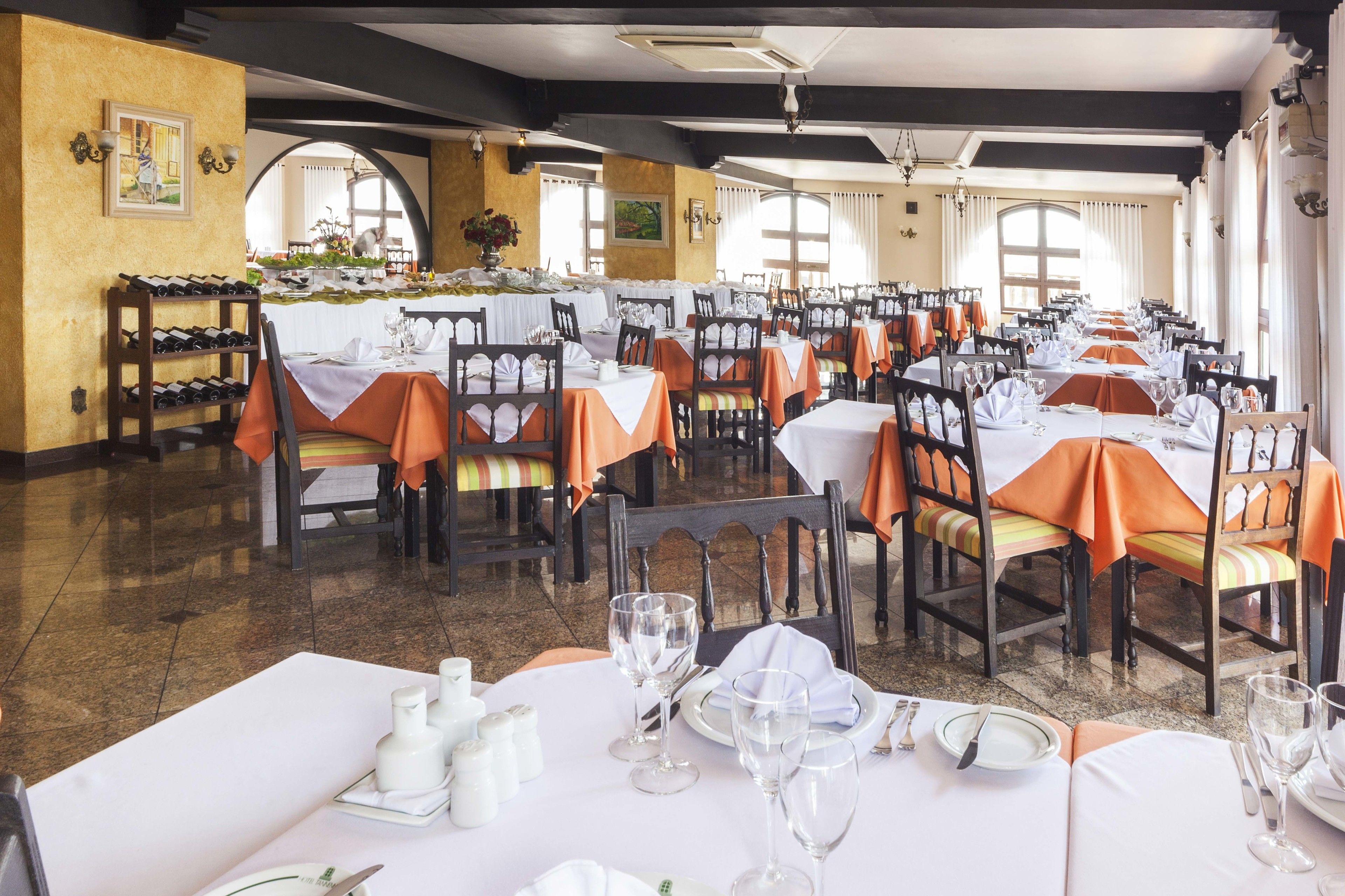 A dining area with tables set for service, featuring a mix of white and orange tablecloths, wooden chairs, and a polished floor.
