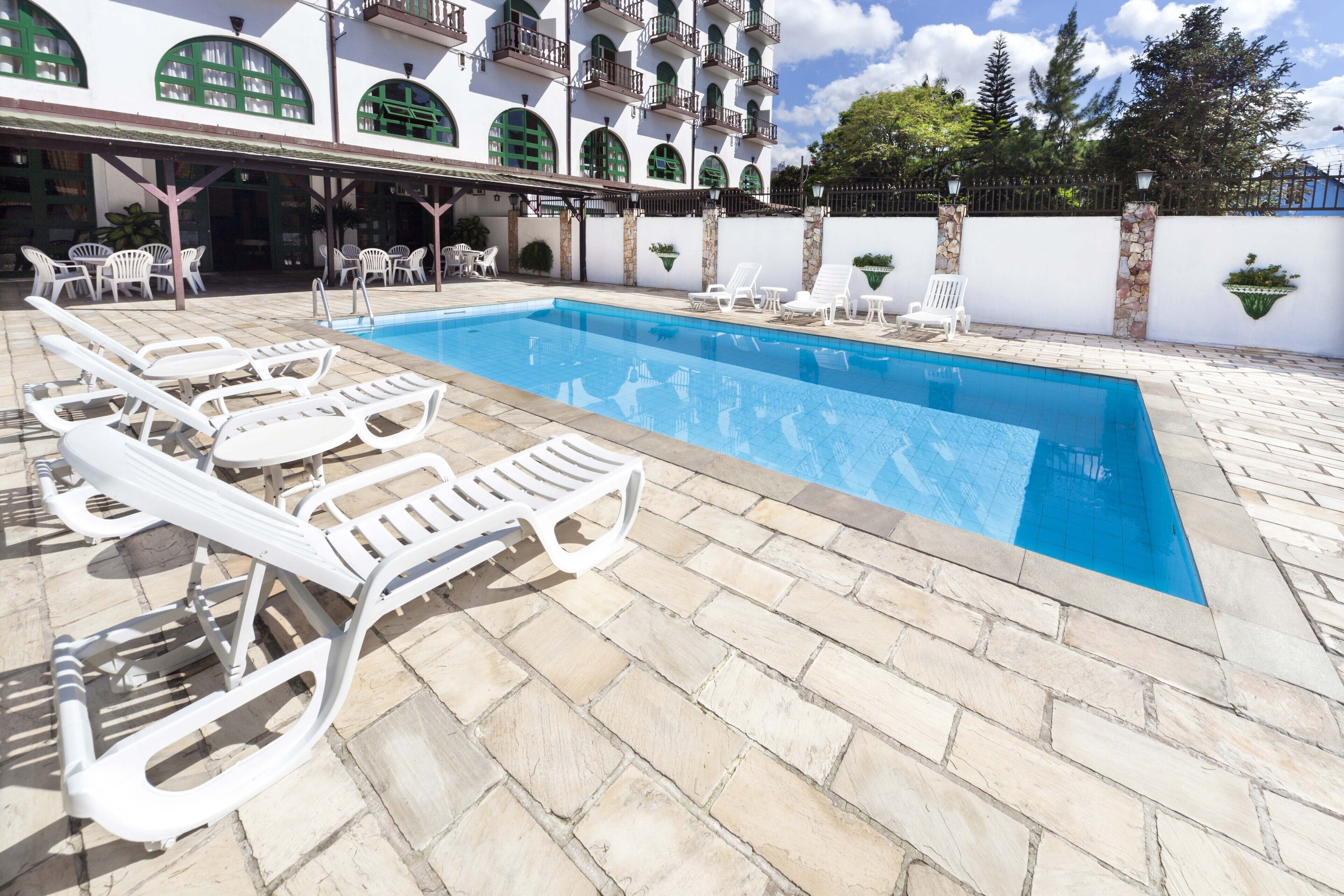 Una piscina con sillones blancos, un deck con baldosas de piedra y un edificio con ventanas y balcones enmarcados en verde.
