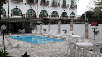 A pool area with lounge chairs and tables, a white building with balconies, and a stone wall.