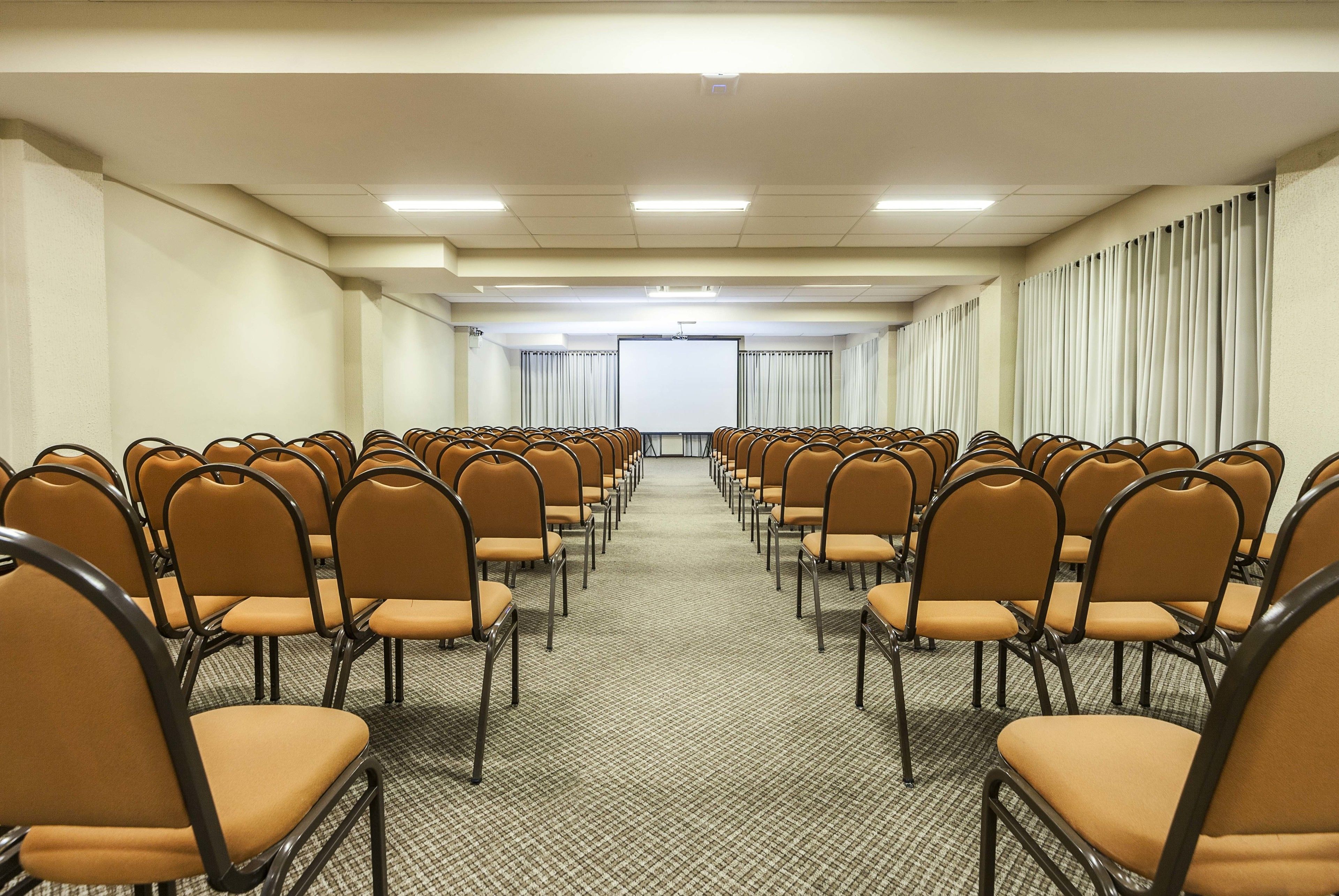 Uma sala de conferências com fileiras de cadeiras voltadas para uma tela de projeção, piso coberto de carpete e cortinas nas janelas.