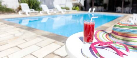 Uma área à beira da piscina com espreguiçadeiras, uma piscina e uma mesa com uma bebida e um chapéu.