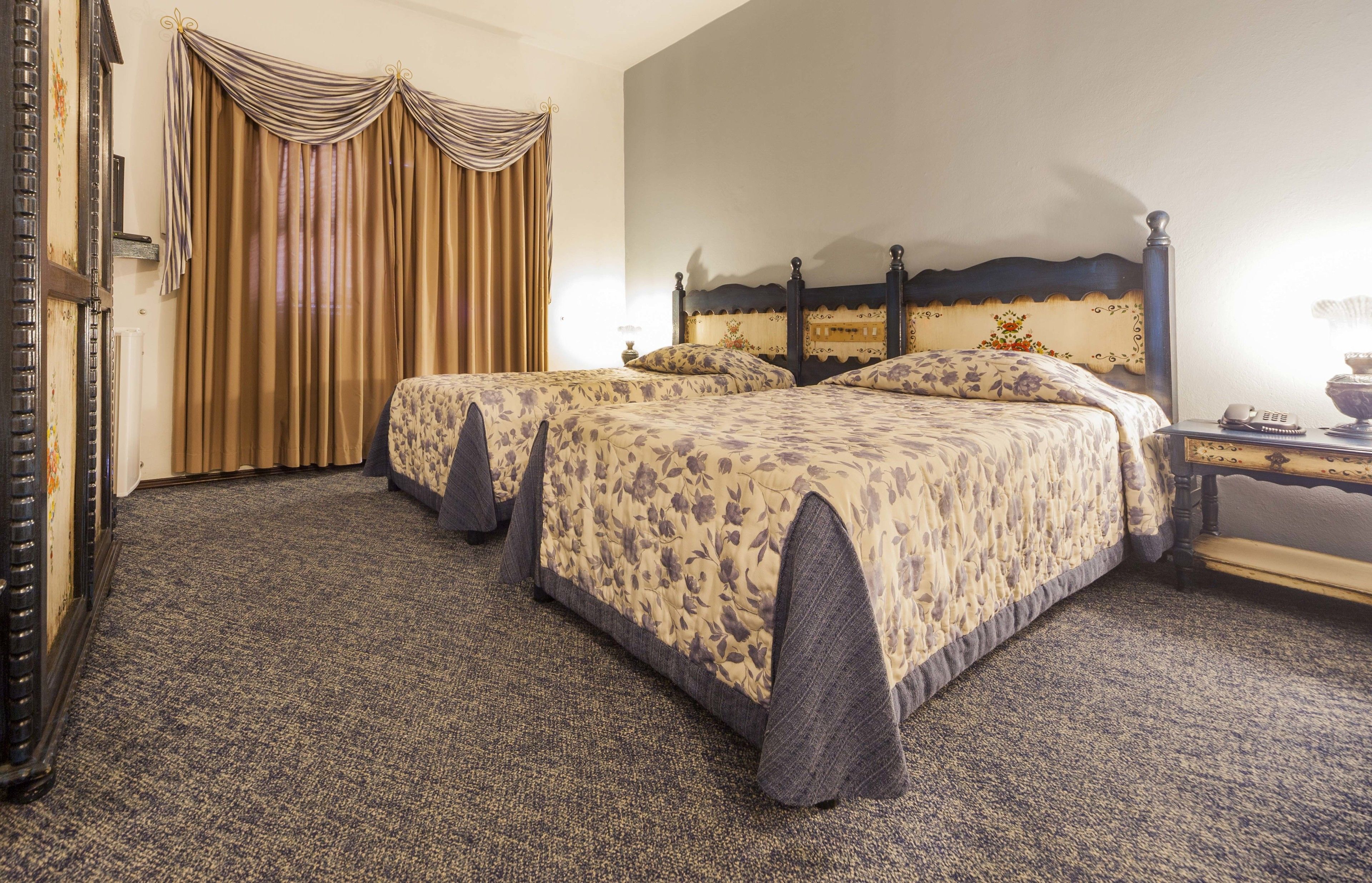 Une chambre d’hôtel avec deux lits, une tête de lit au design classique, un sol recouvert de moquette et une grande fenêtre avec des rideaux.