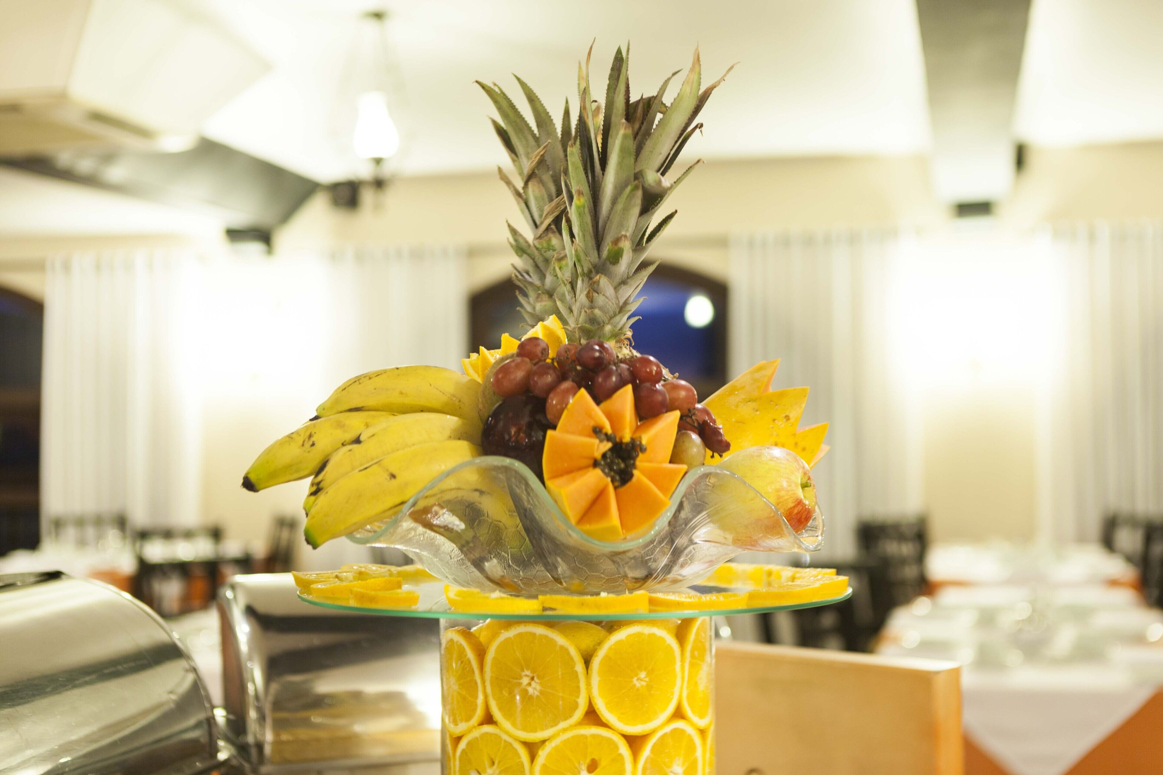 A fruit platter with a pineapple, grapes, and sliced oranges on a glass stand.
