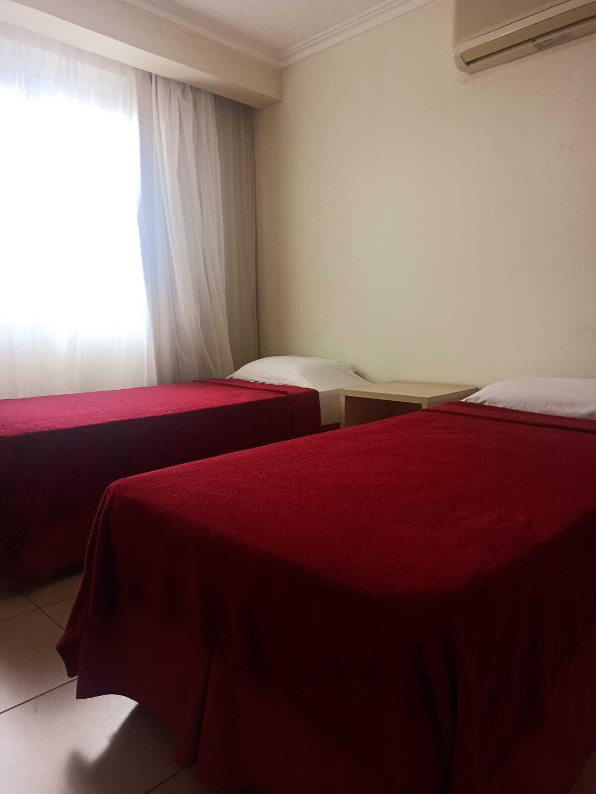 Une chambre d’hôtel avec deux lits simples, une fenêtre avec des rideaux légers et un climatiseur fixé au mur.