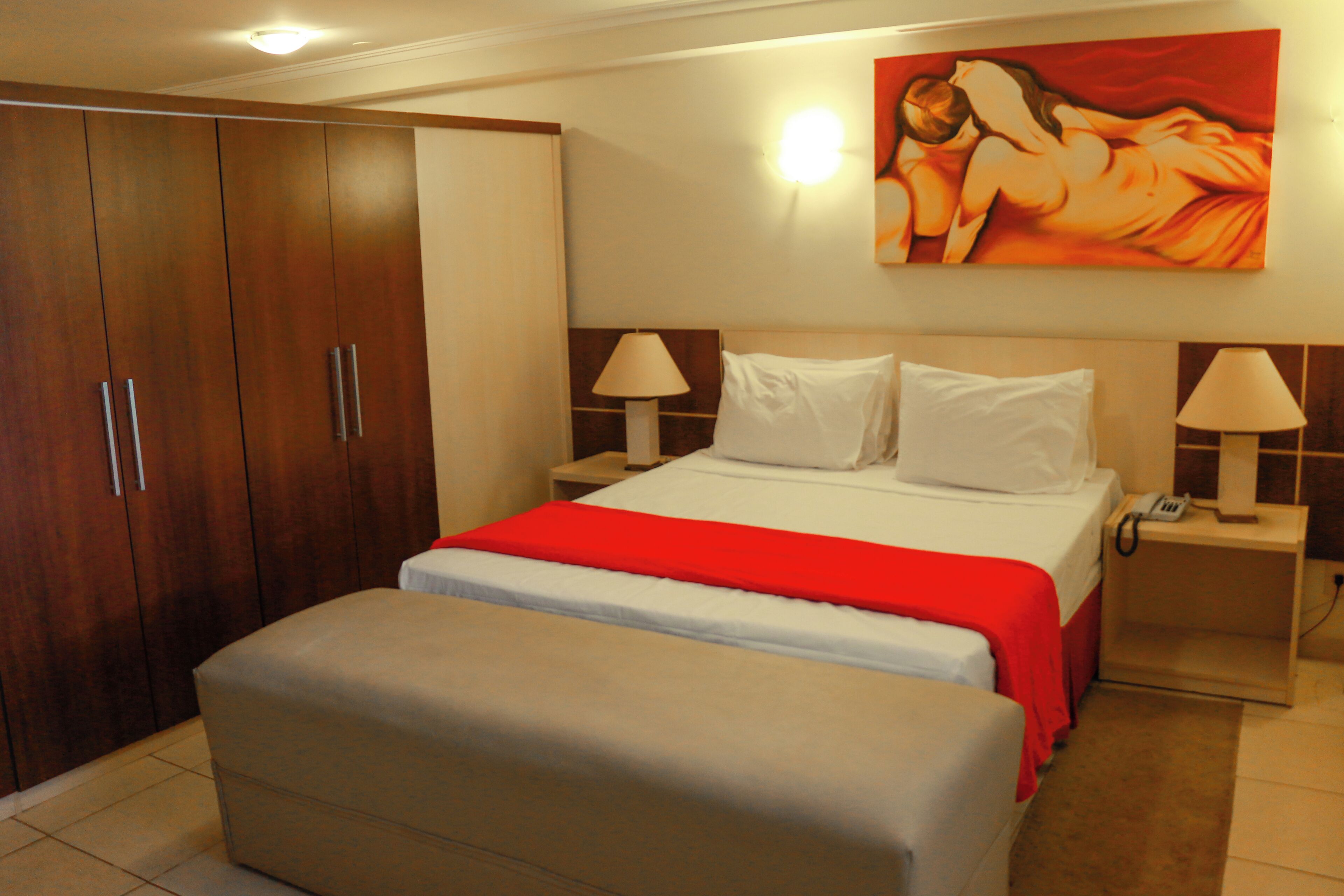 Une chambre d’hôtel avec un lit, des tables de chevet, une couvre-lit rouge, une armoire en bois et un tableau au mur.