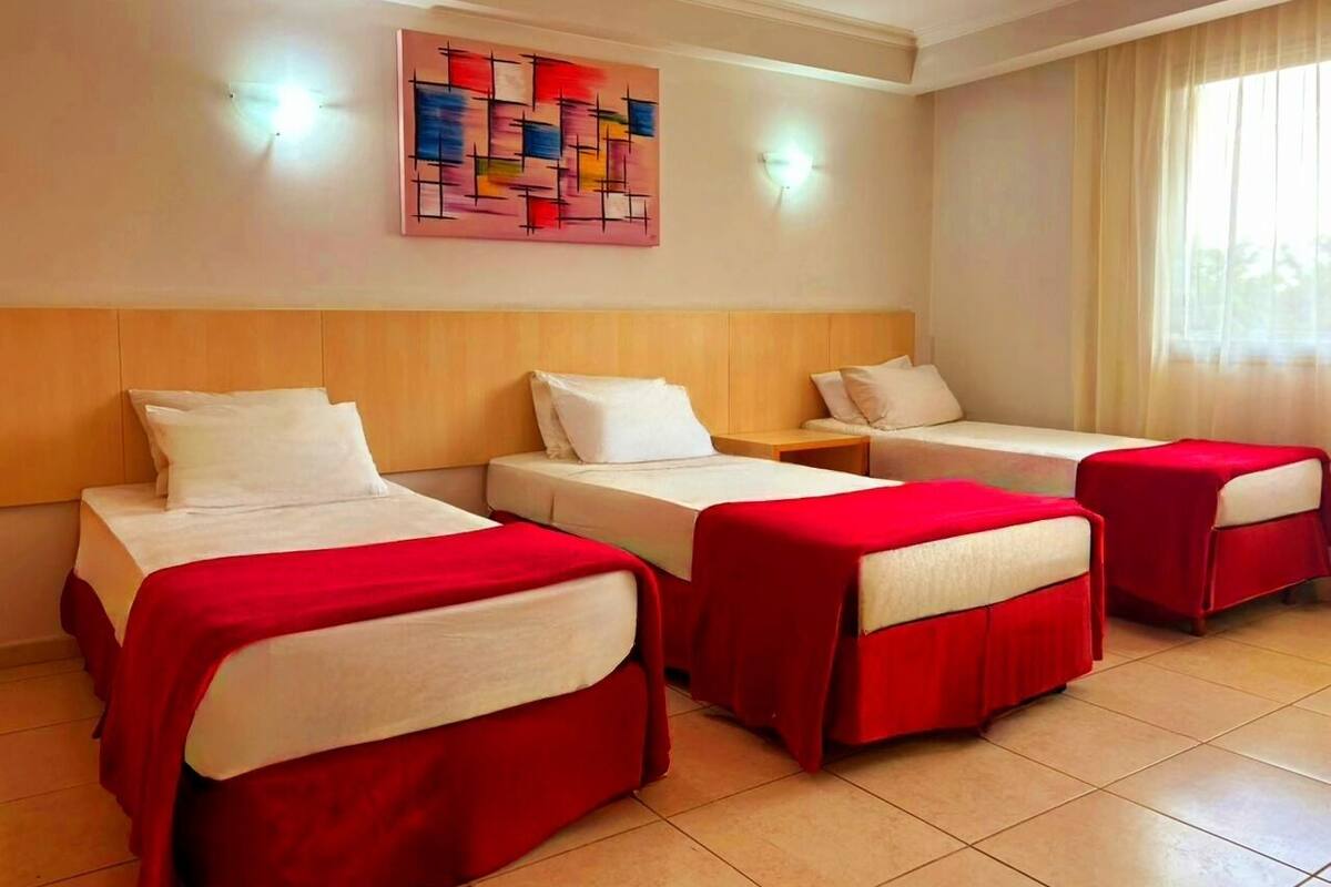 Une chambre d’hôtel avec trois lits, chacun recouvert d’une couverture rouge et agrémenté d’oreillers blancs. Un tableau est accroché au mur au-dessus des lits, et une fenêtre avec des rideaux se trouve à droite.