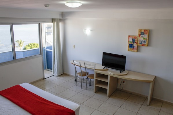 Een hotelkamer met een bed, een bureau met een computerscherm, een stoel en een raam met uitzicht op zee.