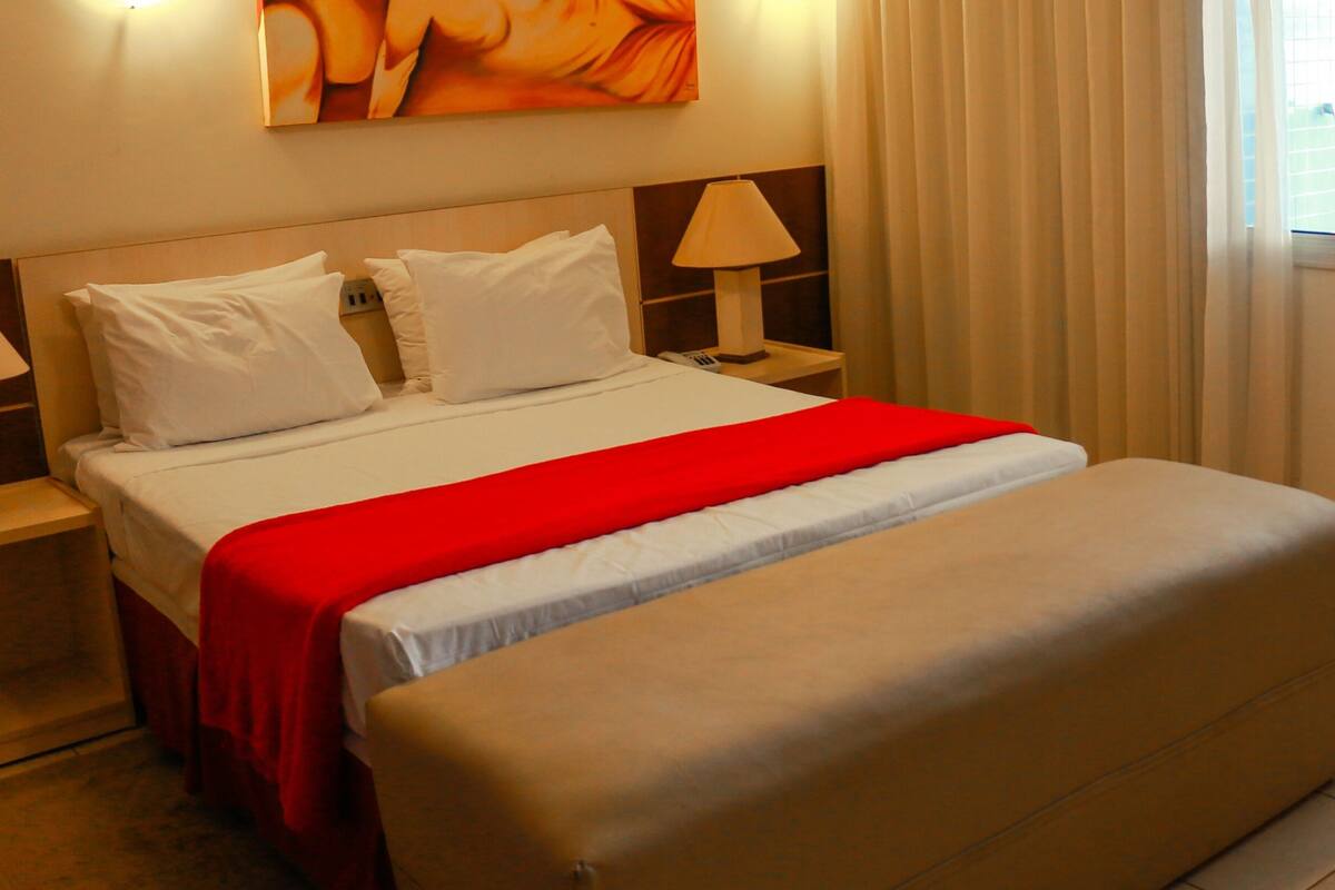 Une chambre d’hôtel avec un grand lit, deux tables de chevet avec des lampes, un plaid rouge et un pouf beige.