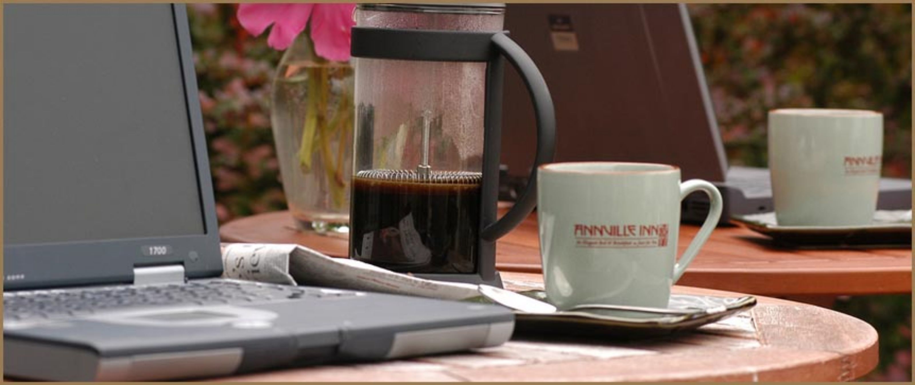 Een laptop, een koffiezetapparaat en twee mokken op een houten tafel.