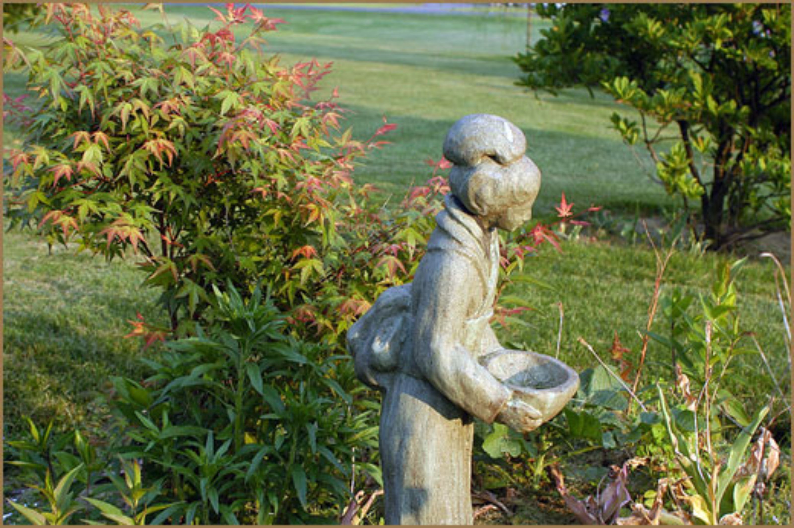 A garden statue of a figure holding a bowl, surrounded by greenery and a lawn.