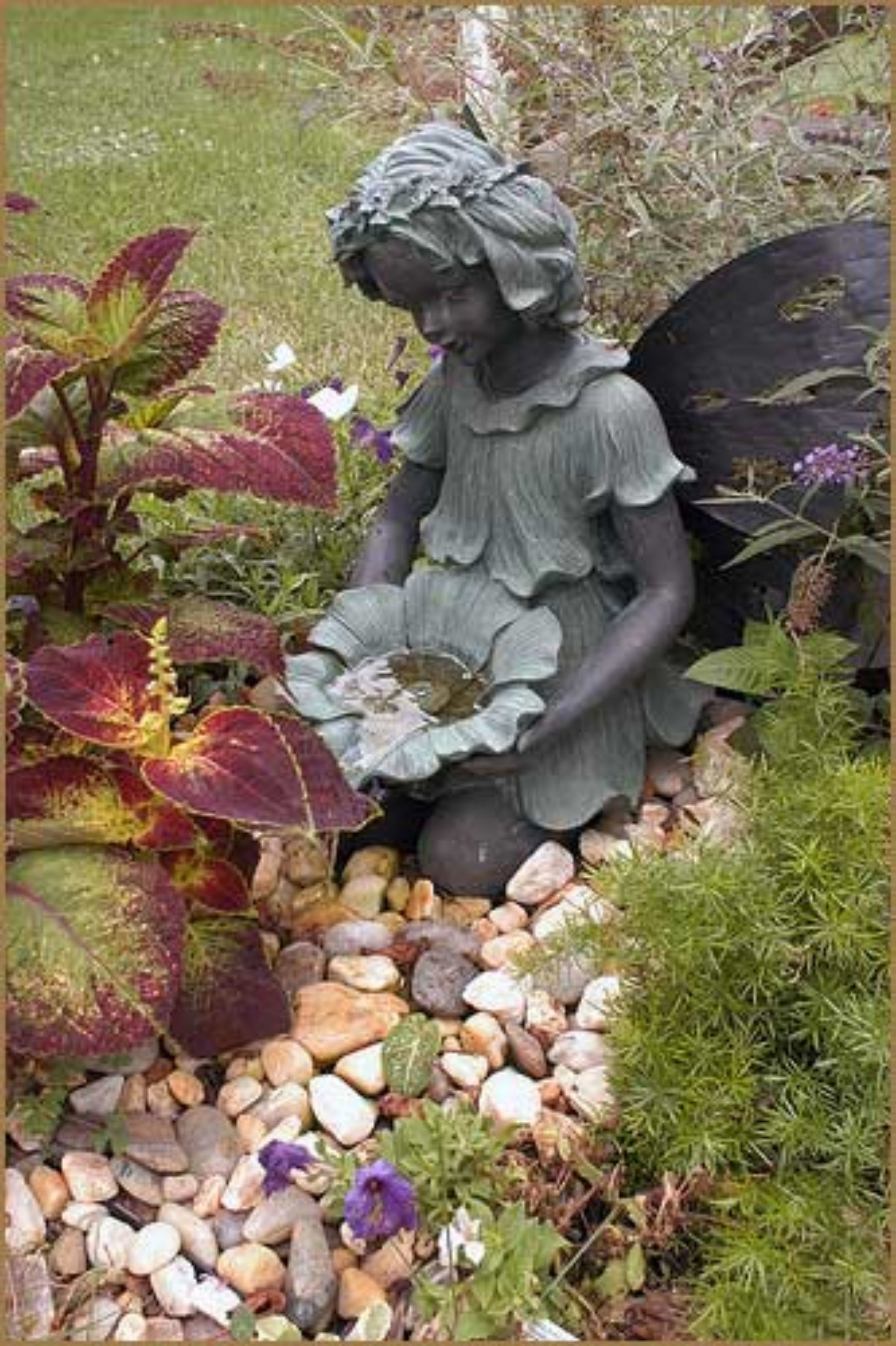 A garden statue of a figure with a ruffled dress and wings, surrounded by various plants and rocks.