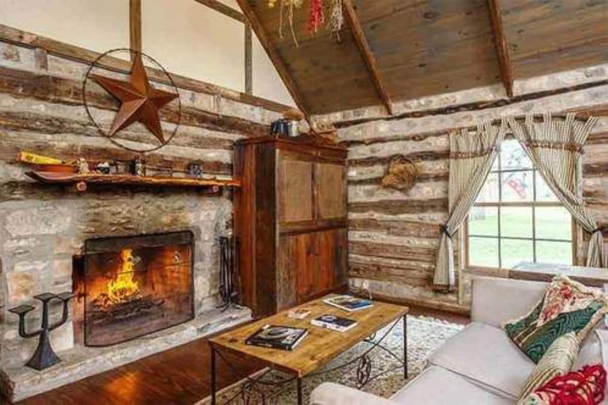 Interior rústico de cabaña con chimenea de piedra, vigas de madera, adorno de estrella y ventana con cortinas.