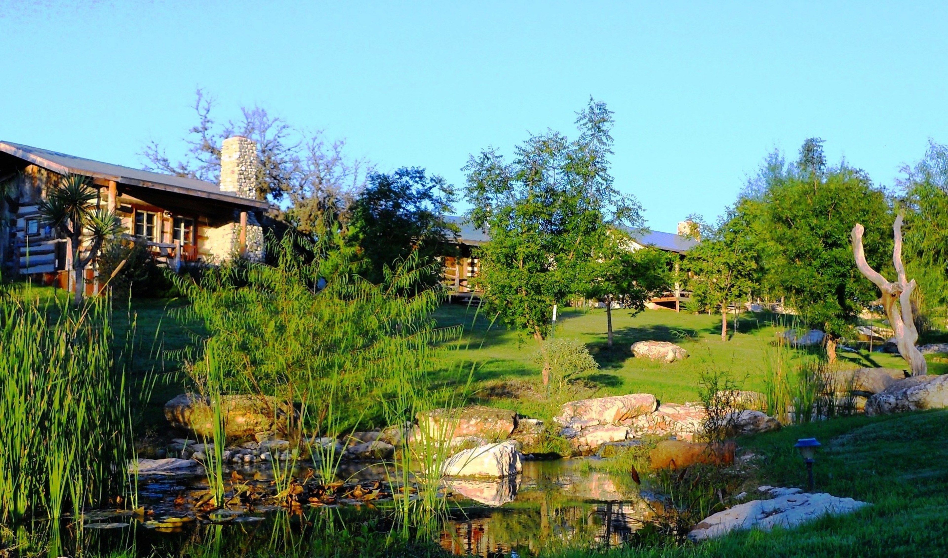 A rustic lodge with a stone chimney, a pond with rocks, and lush greenery.