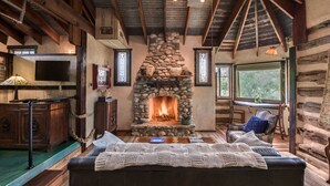 Interior de una cabaña de troncos con chimenea de piedra, muebles de madera y un televisor.
