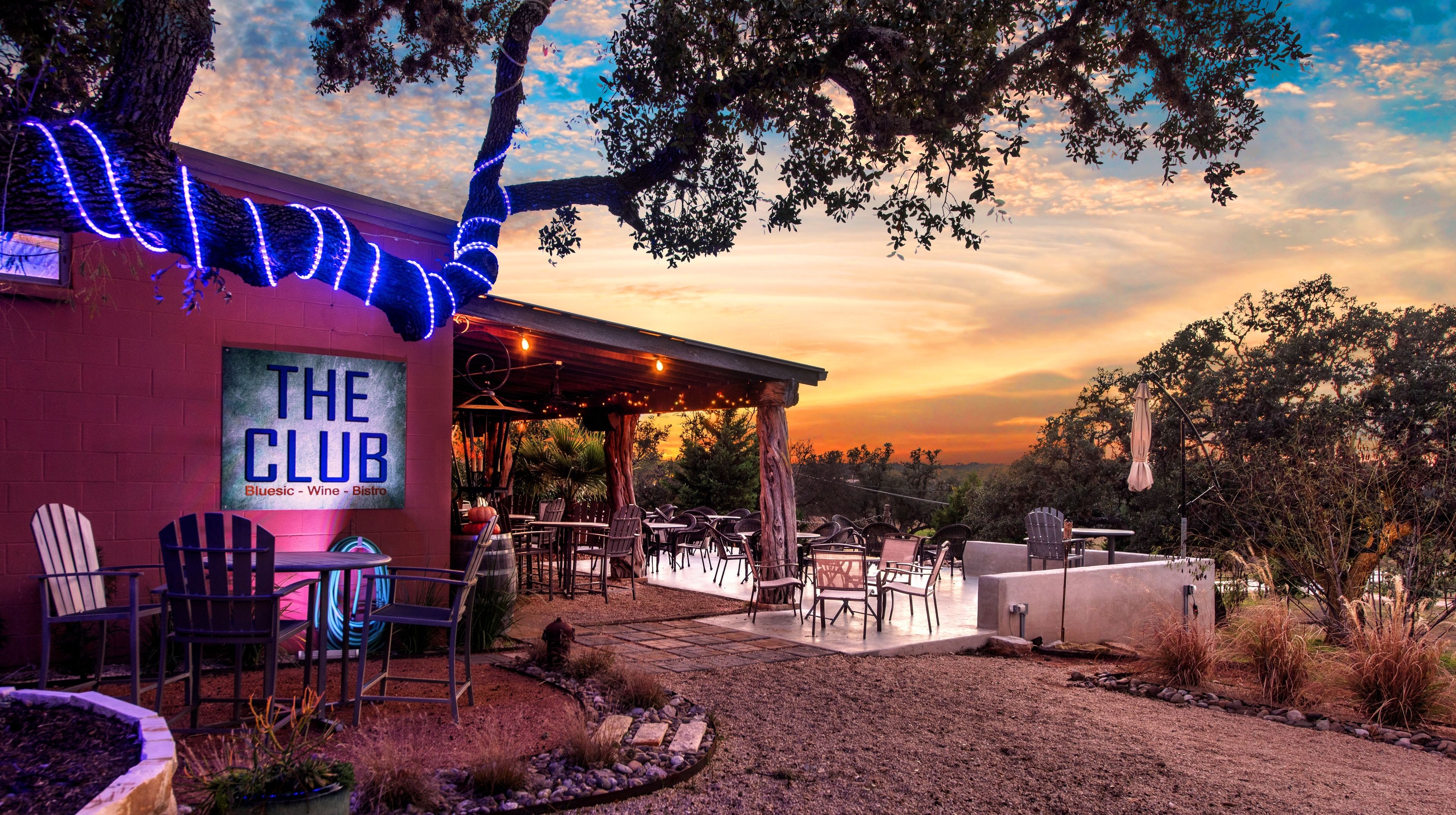 An outdoor seating area with tables and chairs, a sign reading "The Club," and a sunset in the background.