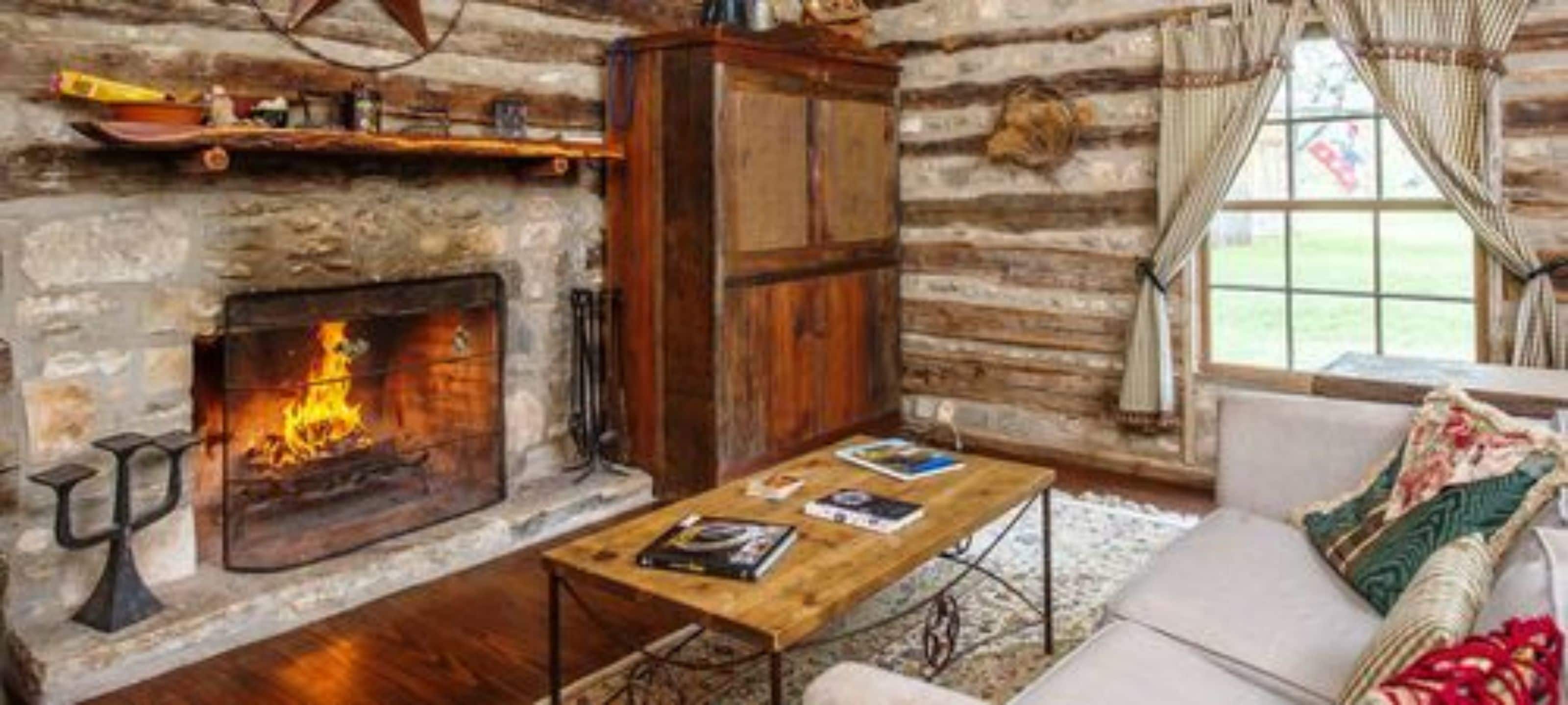A log cabin interior with a stone fireplace, wooden furniture, and a view of the outdoors.