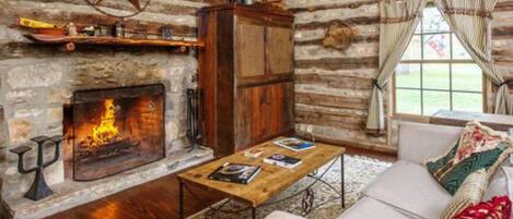 A log cabin interior with a stone fireplace, wooden furniture, and a view of the outdoors.