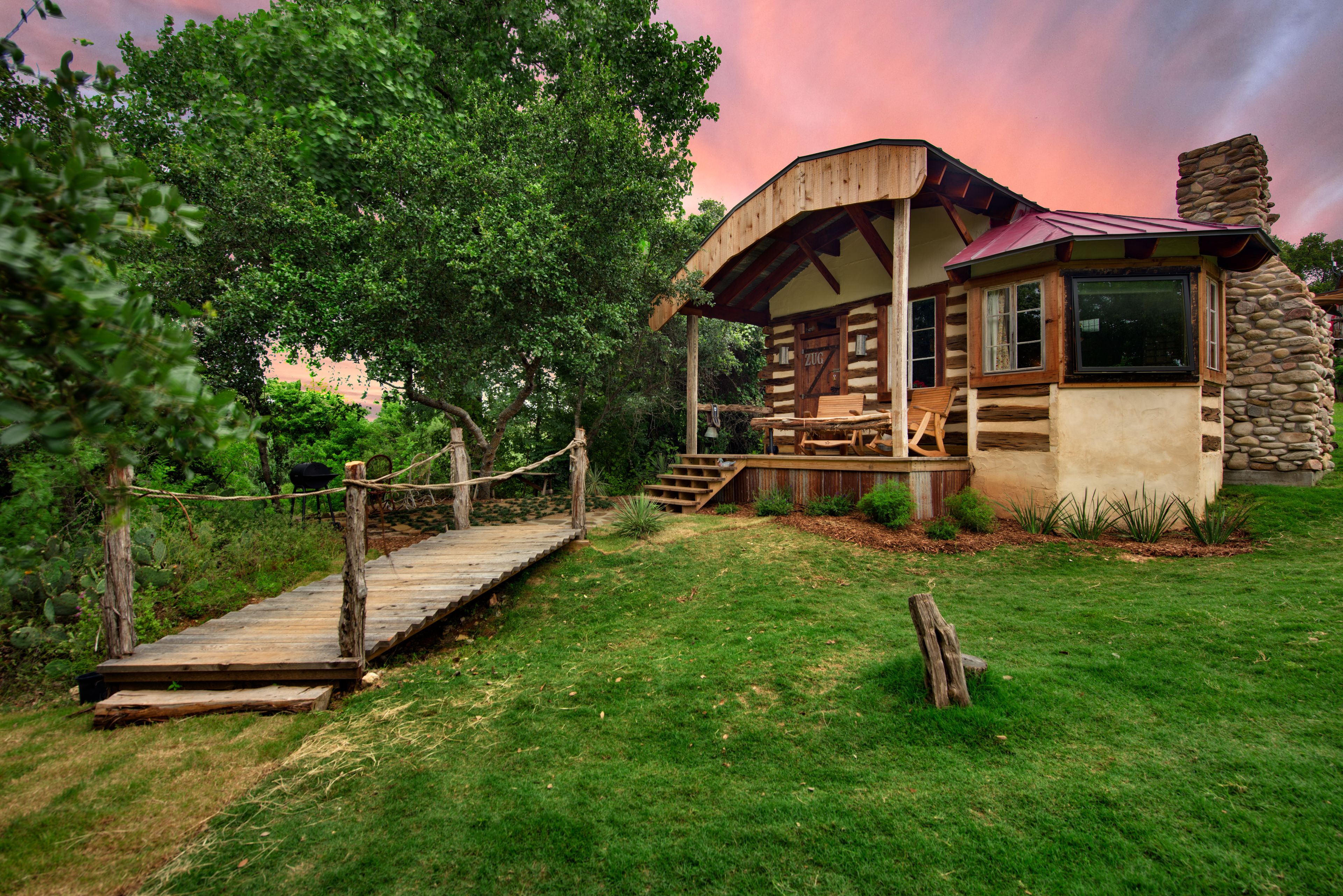 A rustic cabin with a wooden deck and a stone chimney, surrounded by lush greenery.