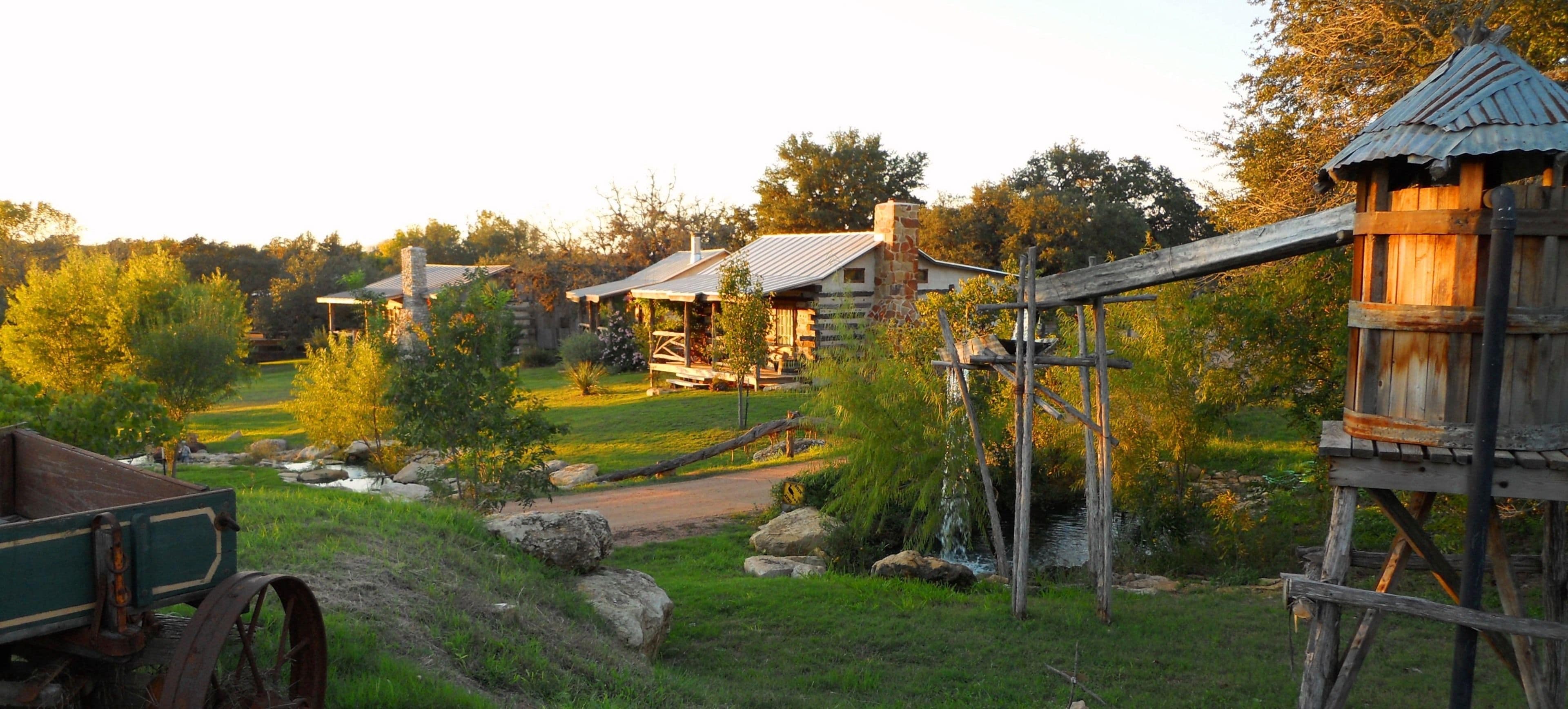 Una cabaña rústica con fachada de madera y techo metálico, rodeada por un paisaje verde exuberante con un pequeño arroyo y un puente de madera.