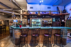 A bar area with a wooden bar counter, three stools, a refrigerator stocked with bottles, and a television mounted on the wall.