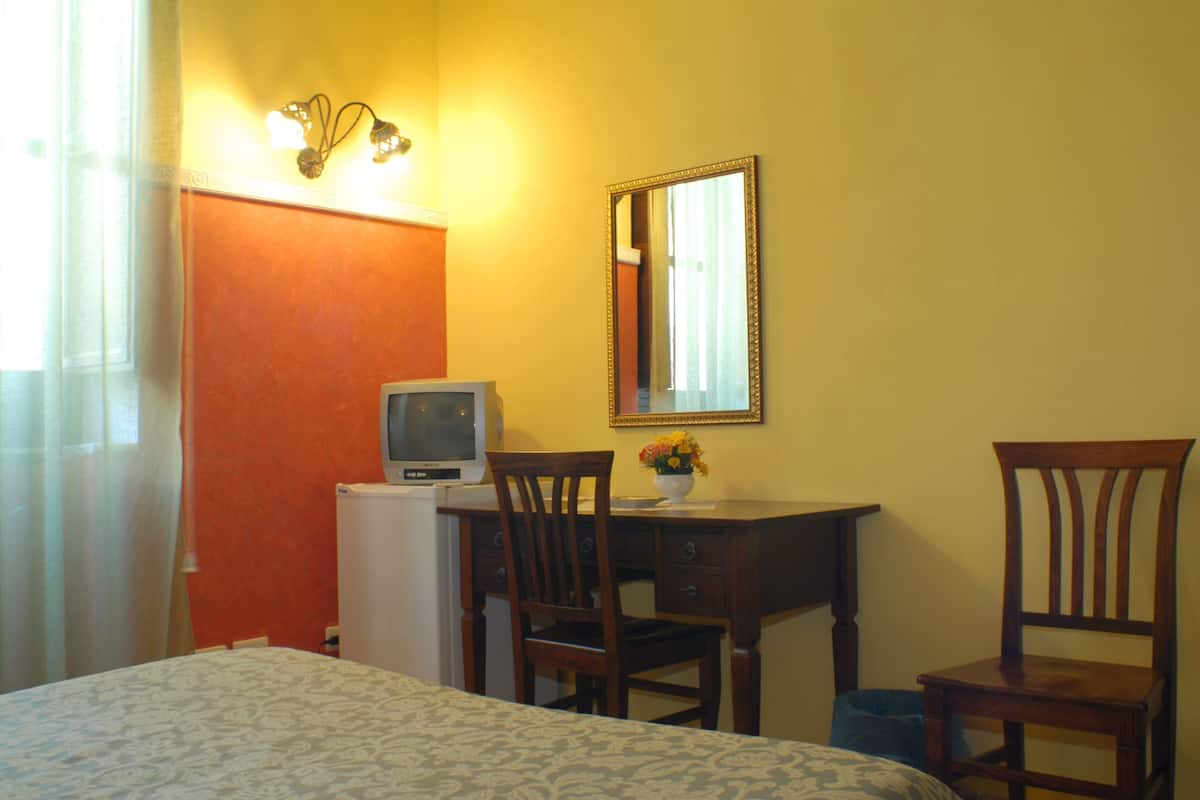 Una habitación de hotel con una cama, un escritorio con computadora, una silla, una mesita con un jarrón de flores y un espejo en la pared.