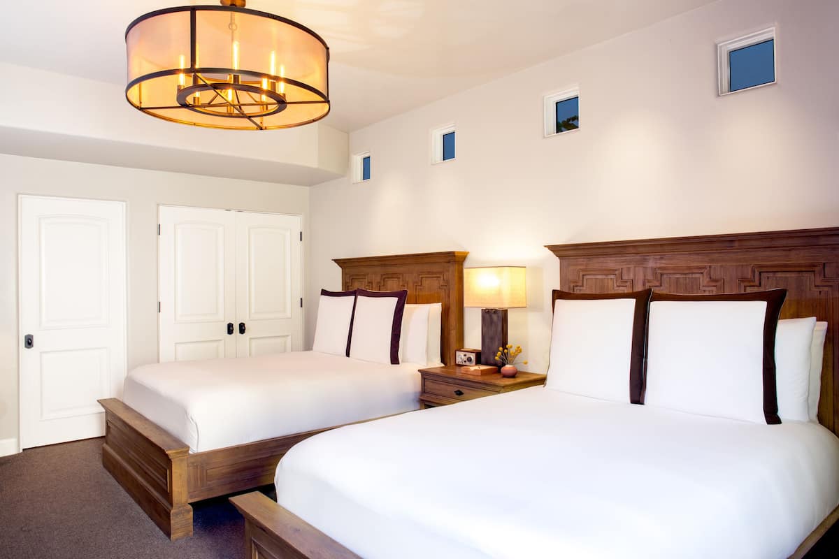 A hotel room with two beds, a wooden headboard, a nightstand with a lamp, and a ceiling light fixture.
