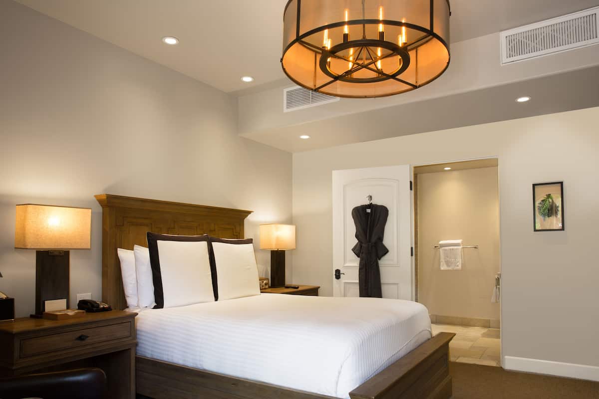 A hotel room with a large bed, two bedside tables with lamps, a wooden headboard, a robe hanging on the door, and a framed picture on the wall.