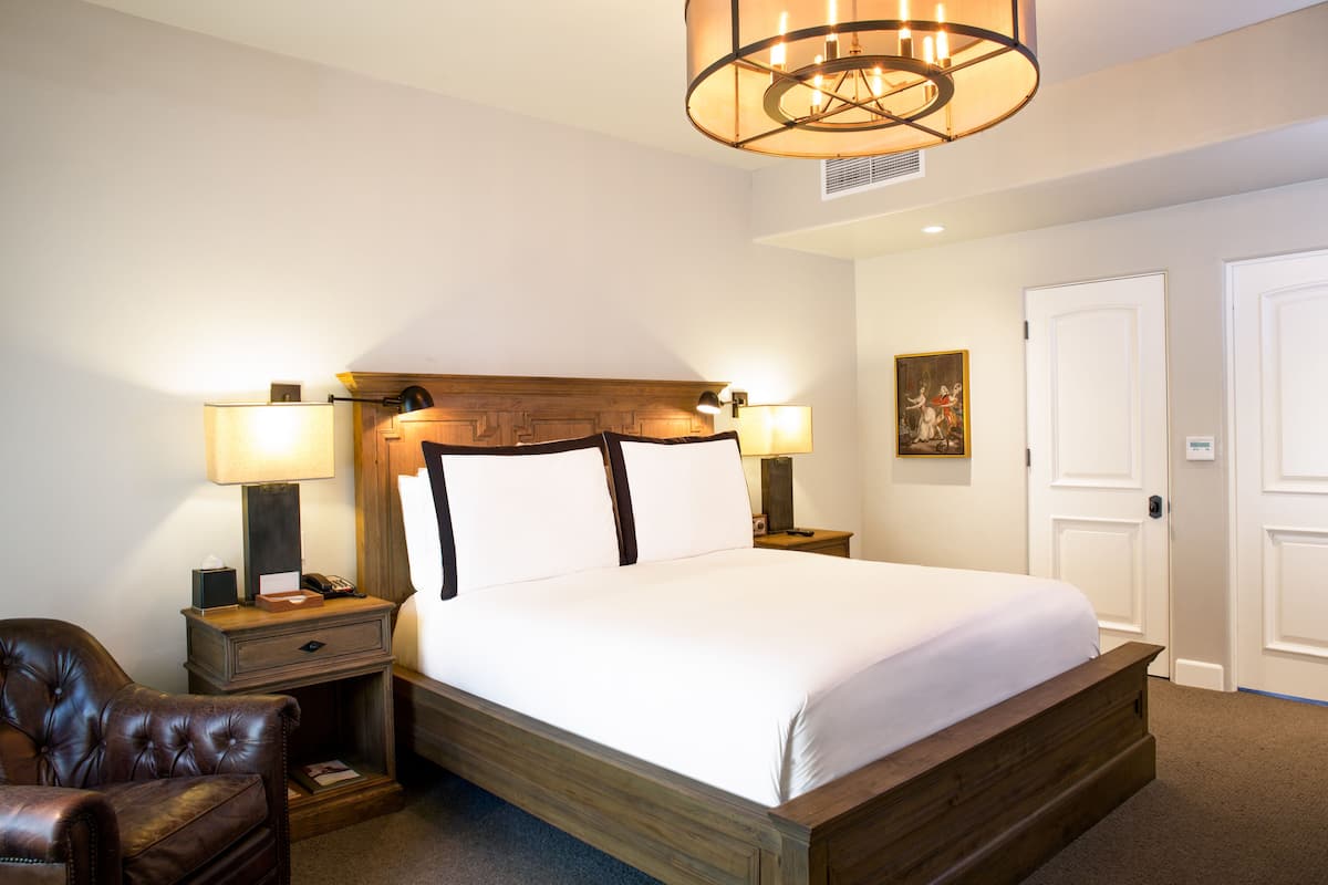 A hotel room with a large bed, two bedside tables with lamps, a leather armchair, and a wooden headboard.