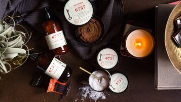 A variety of personal care products including body scrub, hand cream, and shampoo, arranged on a textured surface with a candle and decorative items.