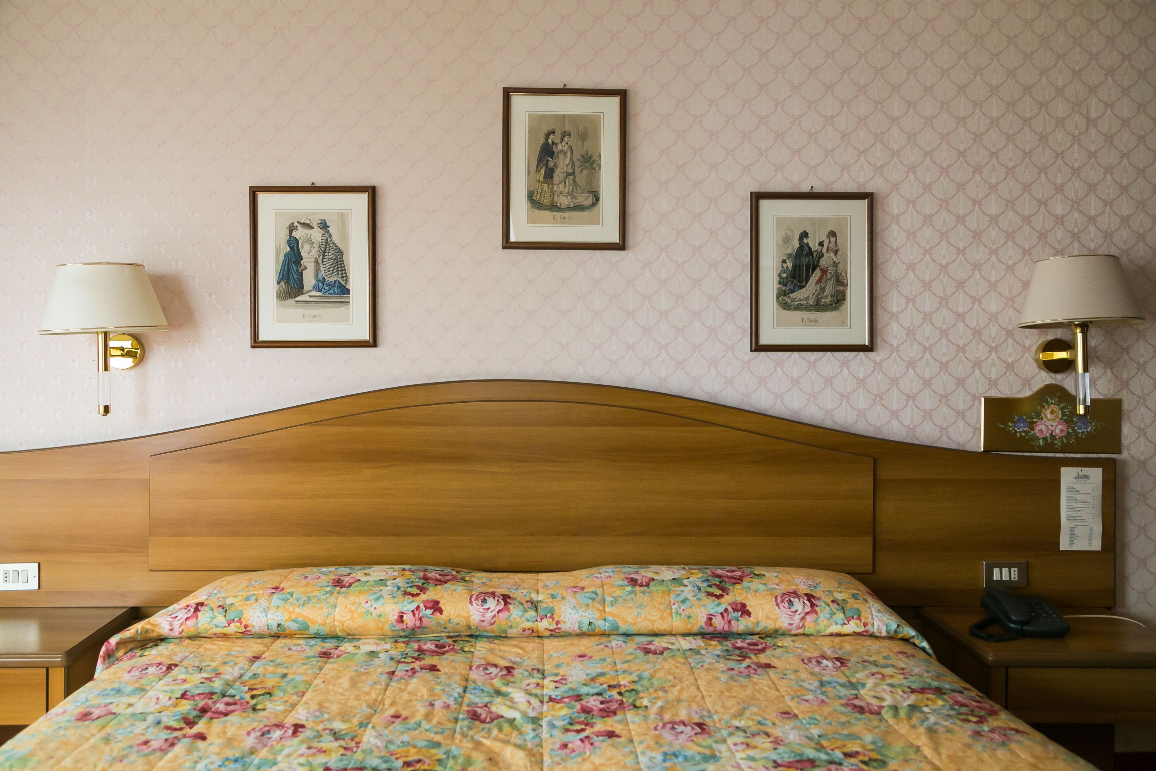 Habitación de hotel con cabecera de madera, ropa de cama floral, dos lámparas de noche y tres cuadros en la pared.