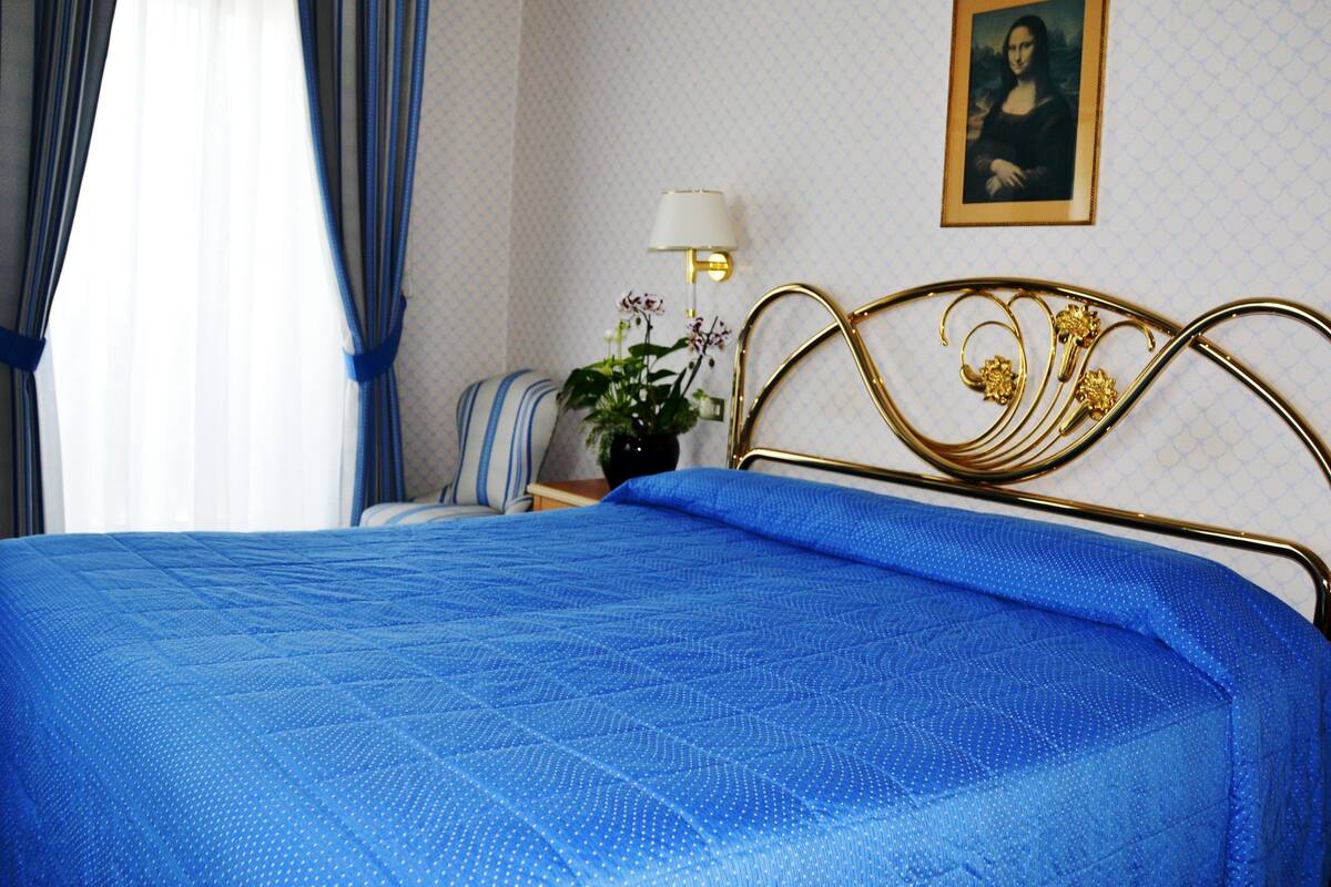 Un lit avec une couette bleue, une tête de lit ornée d’un cadre doré, une plante en pot, une lampe et une œuvre d’art encadrée accrochée au mur.