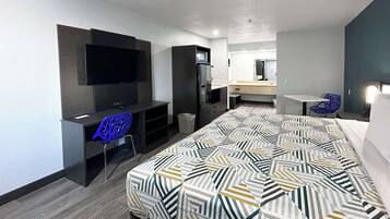 Habitación de hotel moderna con una cama grande, un escritorio con silla, un televisor montado en la pared y una pequeña cocina.