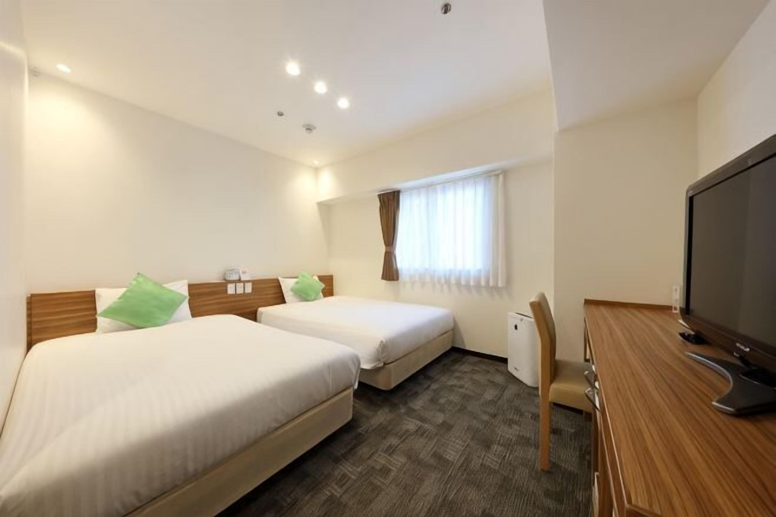 Quarto de hotel com duas camas, uma escrivaninha, televisão e janela com cortinas.