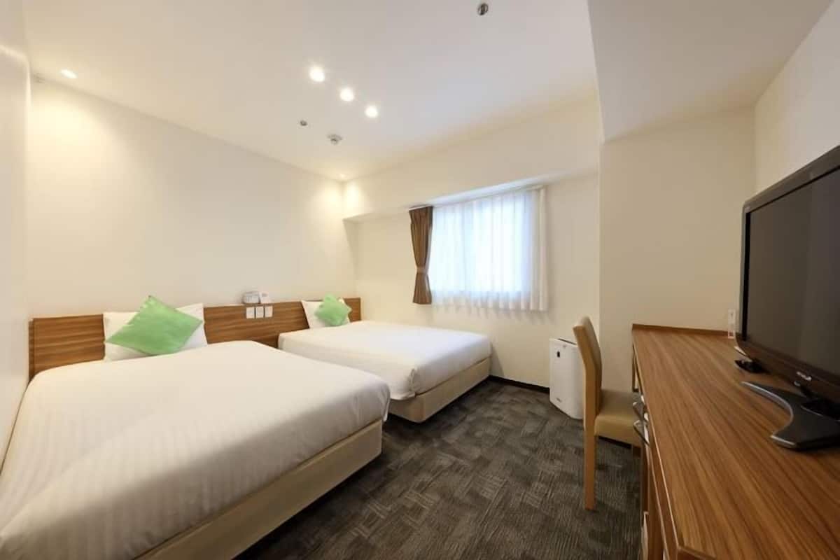 Quarto de hotel com duas camas, uma escrivaninha, televisão e janela com cortinas.