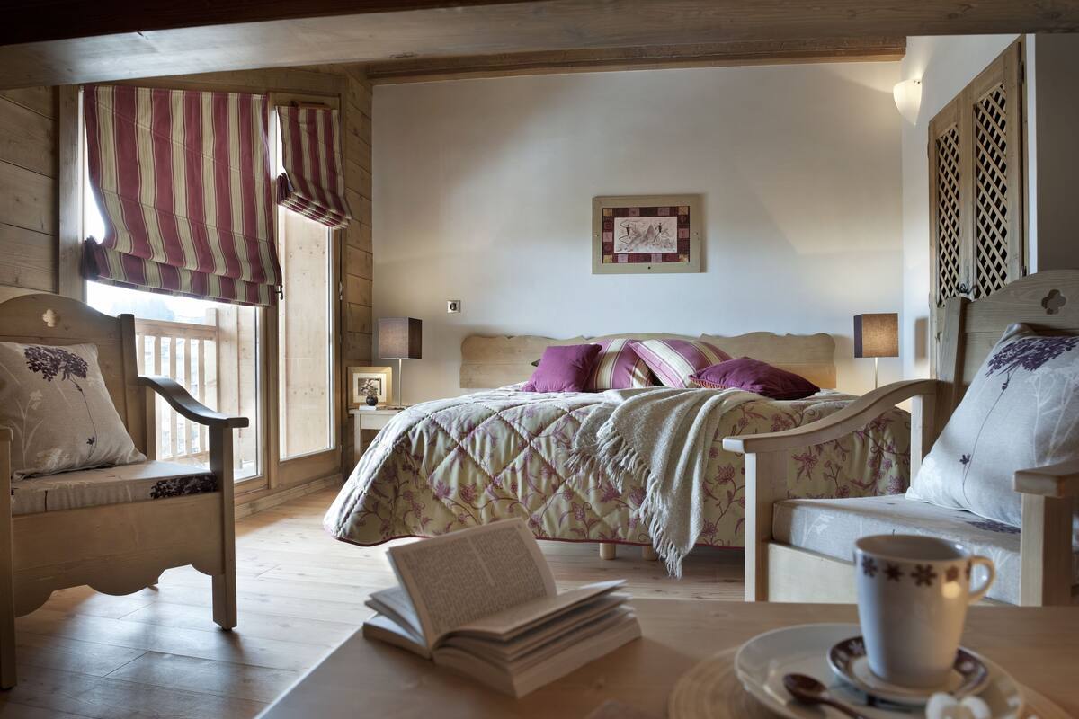 A cozy bedroom with a bed, two chairs, a table with a cup and saucer, and a window with curtains.