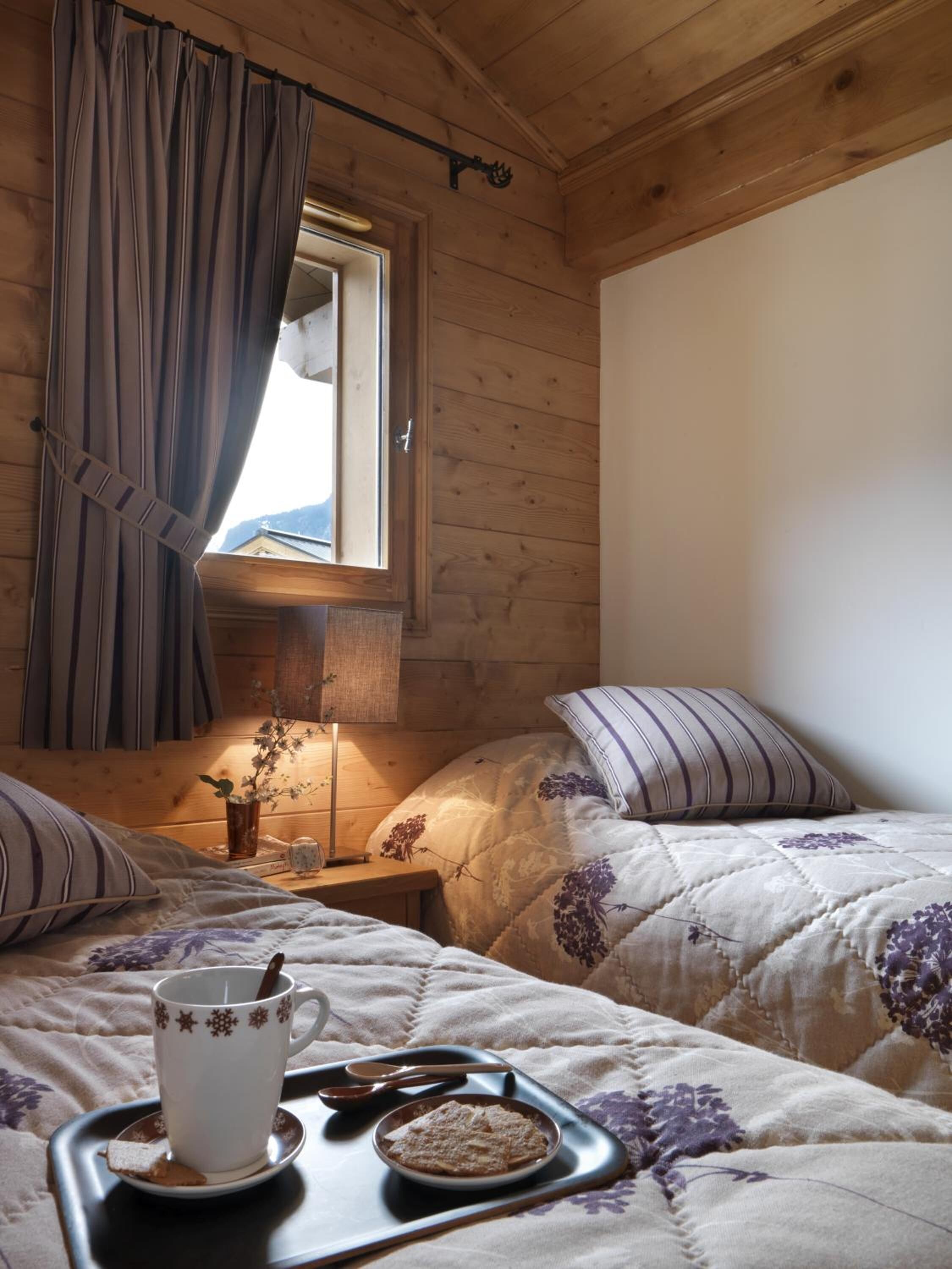 A cozy room with wooden walls and a window with curtains. There is a bed with a floral quilt, a bedside table with a lamp, and a tray with a cup and a cookie.
