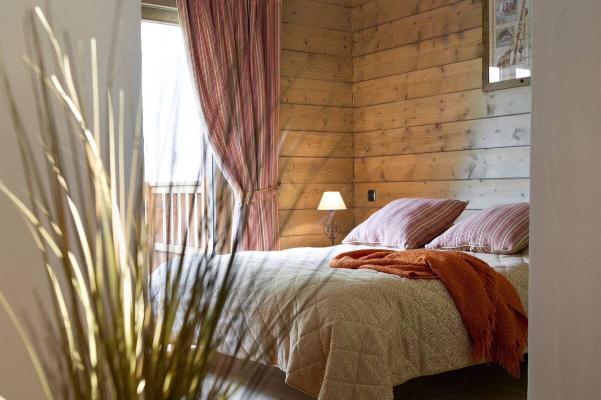 Un dormitorio acogedor con techo de madera, una cama con almohadas a rayas, una lámpara de noche y una ventana con cortinas.