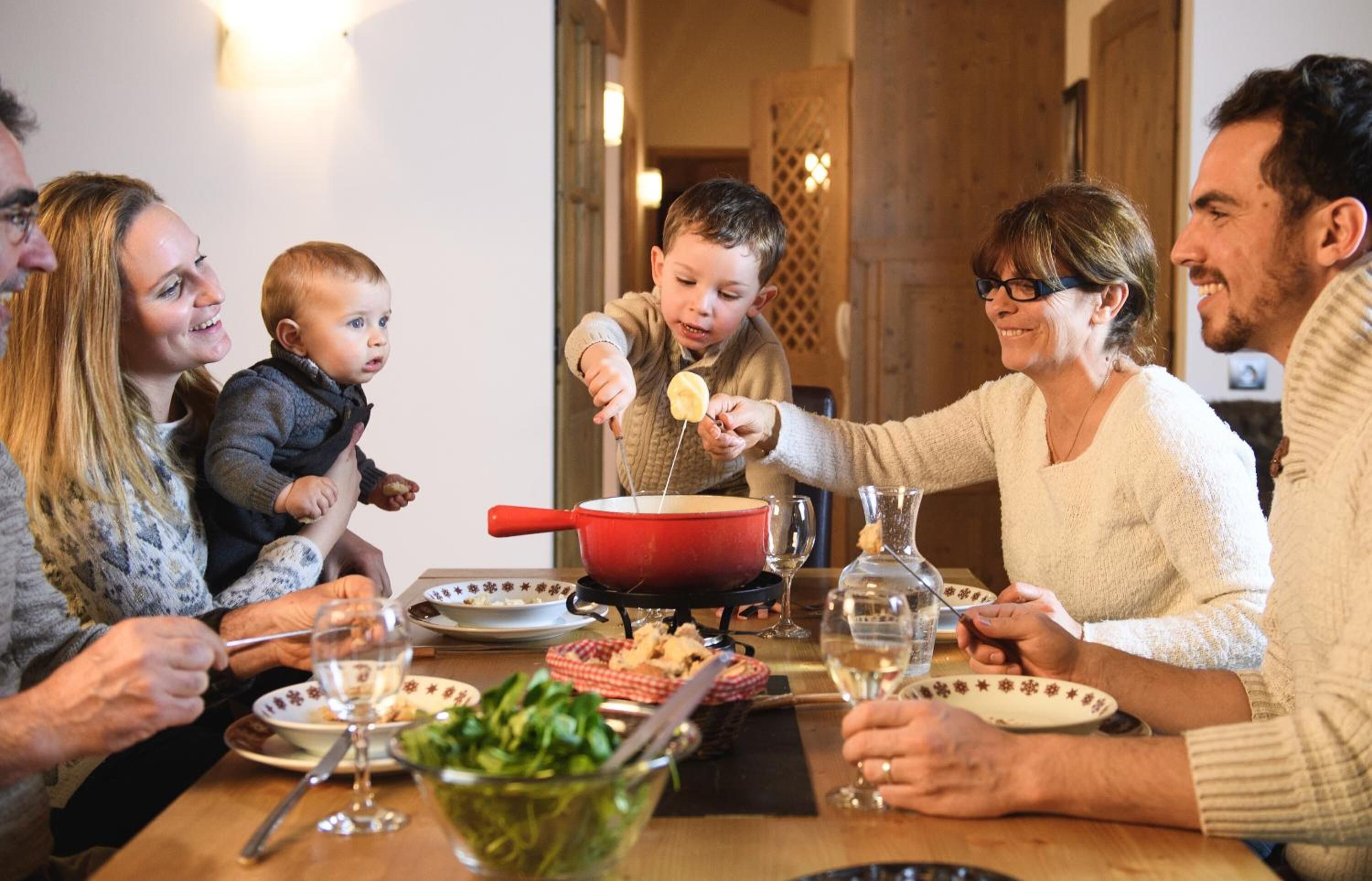 Eine Familie sitzt an einem Holztisch und isst, mit einem roten Topf, Tellern und Gläsern.