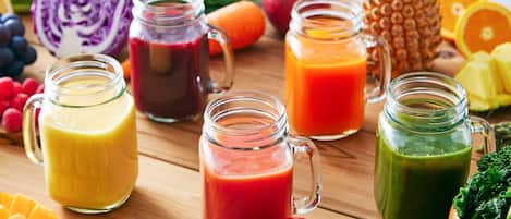 A wooden table with various fruits, vegetables, and three glass jars containing different colored smoothies.