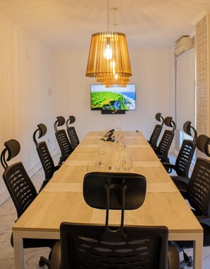 Une salle de conférence avec une longue table, des chaises noires, un téléviseur fixé au mur et un lampadaire suspendu.