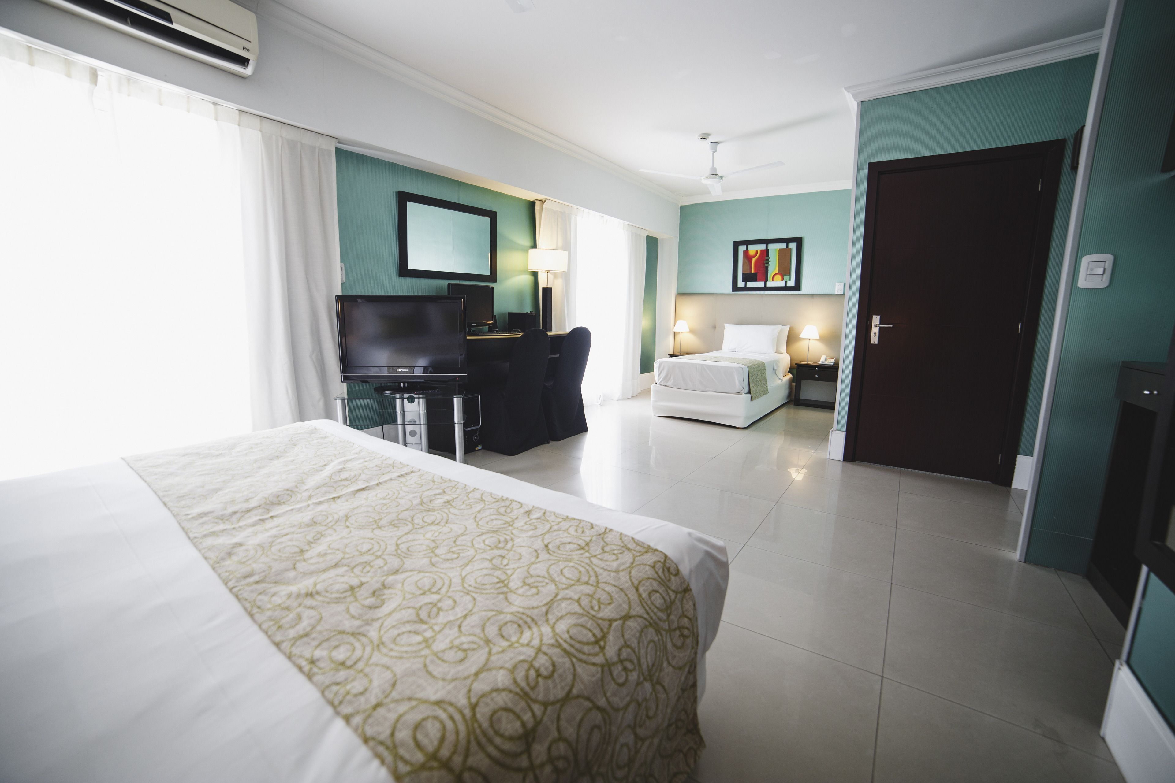 Habitación de hotel con una cama grande, un escritorio con computadora, un televisor y una puerta que da a otra habitación.