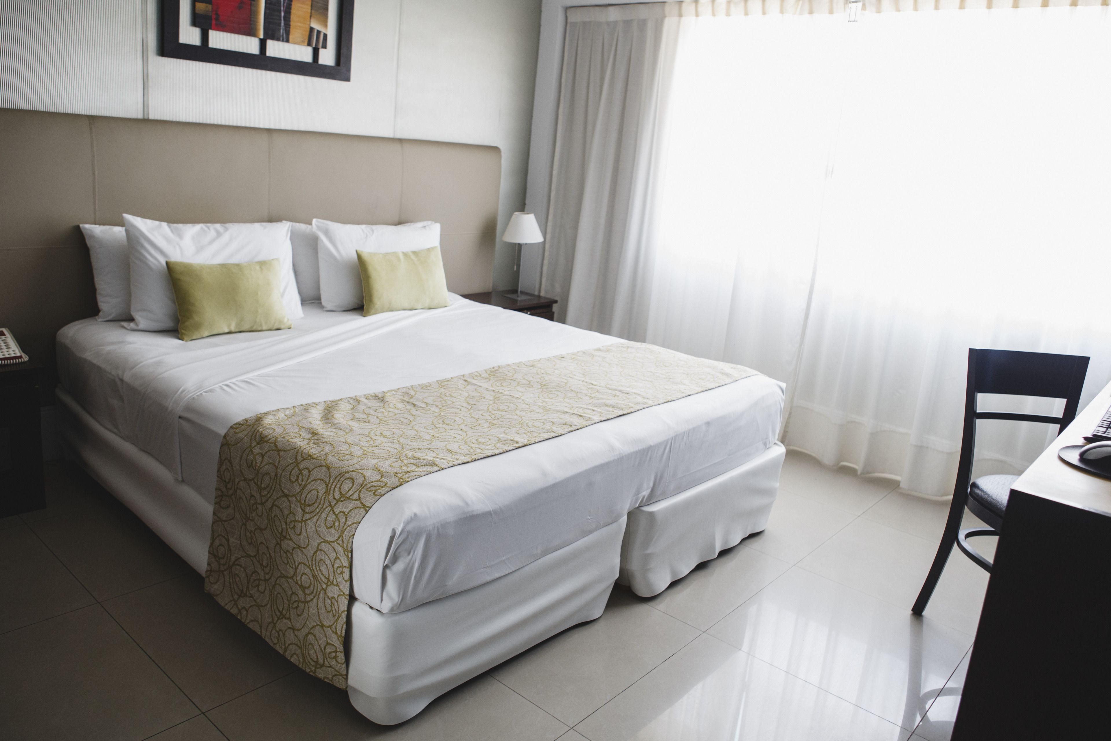 A neatly made bed with a patterned bedspread, a bedside table with a lamp, and a chair in a bright room with a large window.
