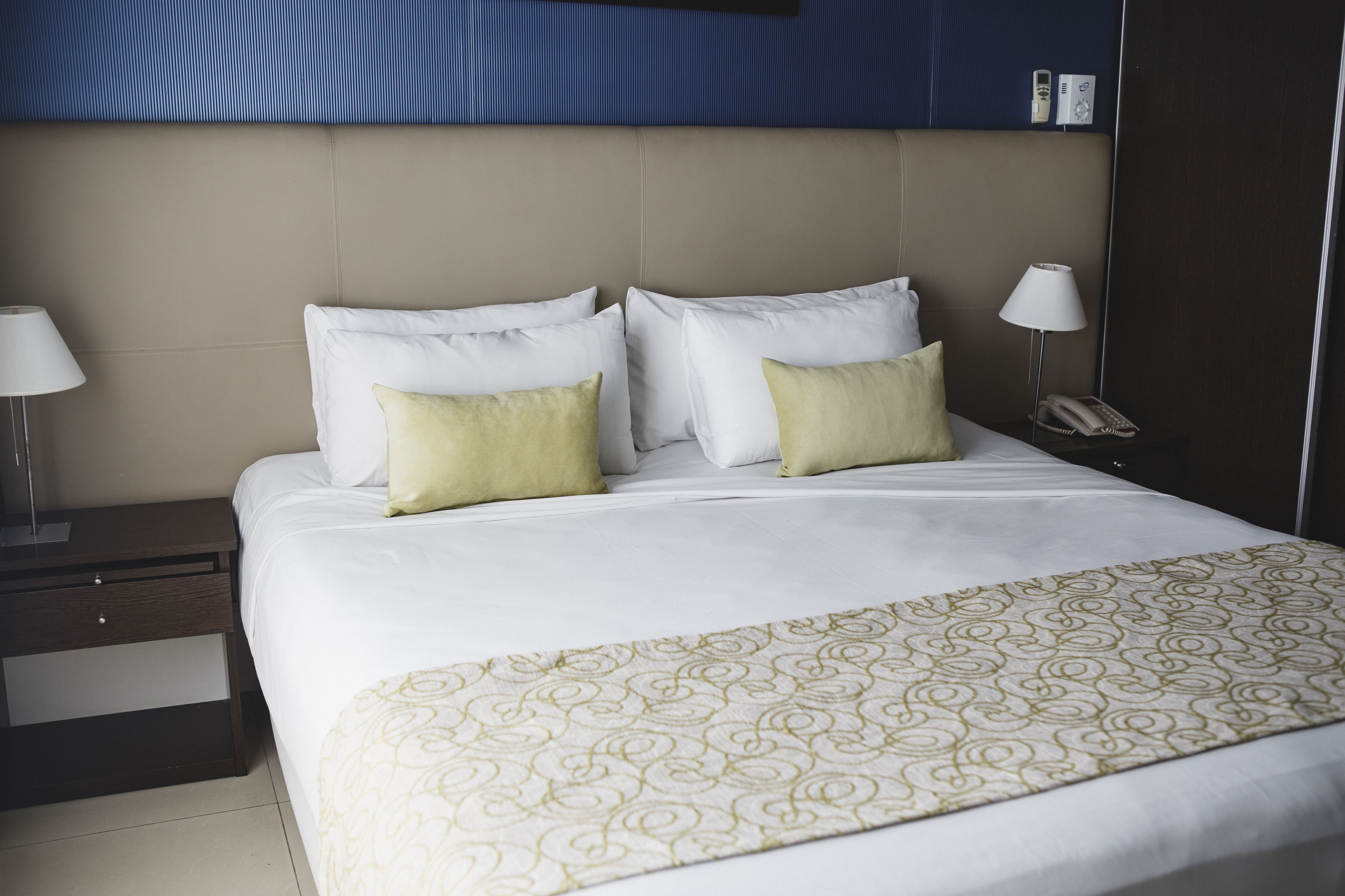 A neatly made bed with white and green pillows, a patterned throw blanket, and a bedside table with a lamp and a phone.