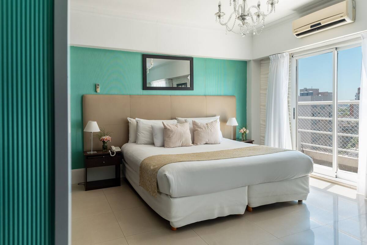 Une chambre d’hôtel moderne avec un grand lit, une table de chevet avec une lampe, un miroir et une vue sur la ville par une grande fenêtre.