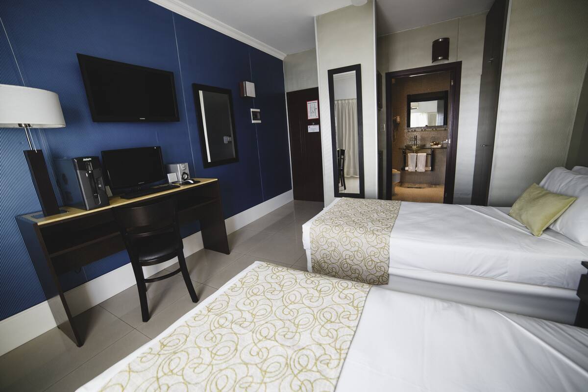 Une chambre d’hôtel équipée d’un bureau, de deux lits, d’une télévision et d’une salle de bain visible à travers une porte ouverte.
