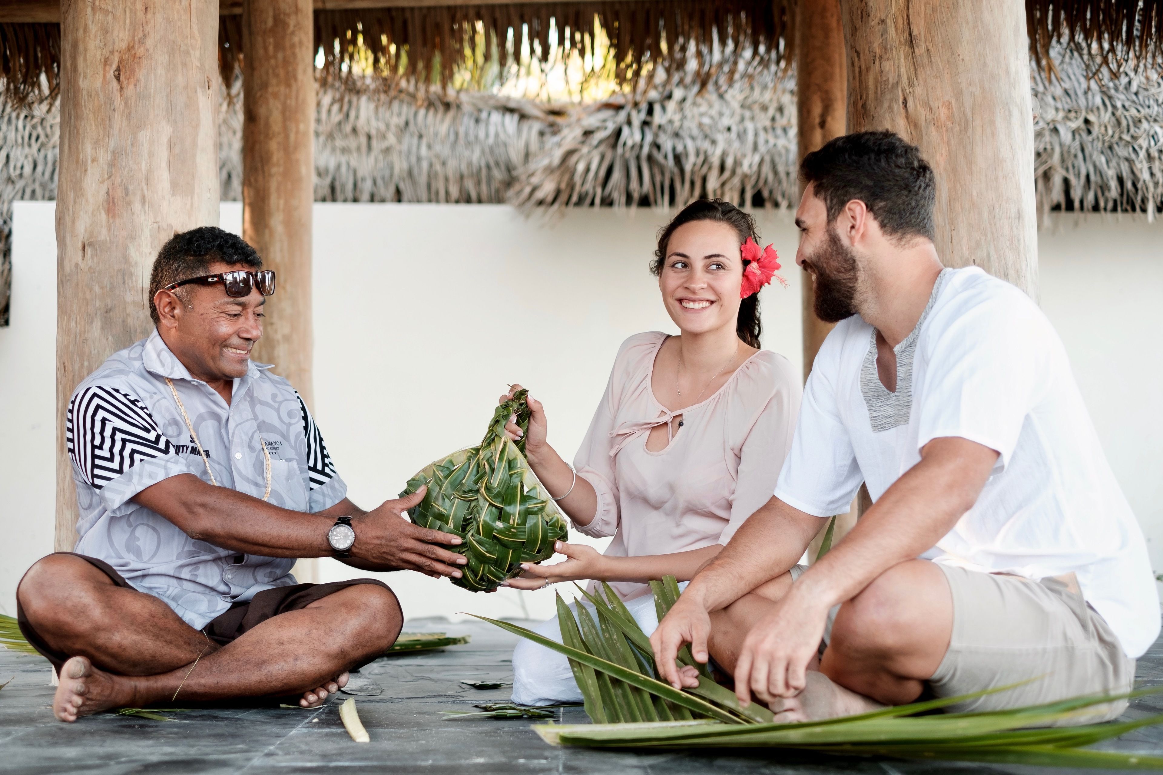 Three people sitting on a tiled floor, exchanging a woven basket.