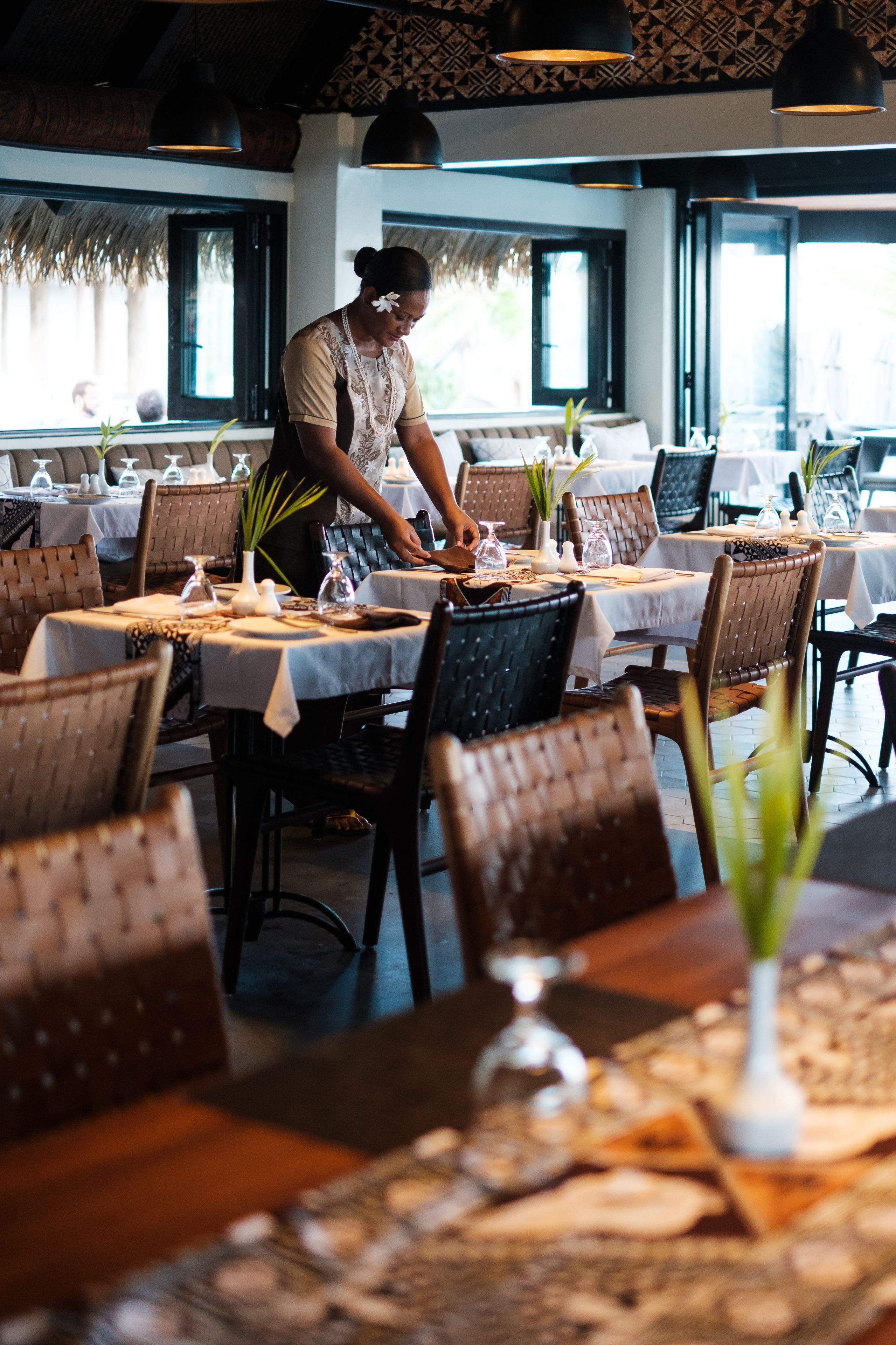 Un restaurant avec des tables dressées pour le service, un serveur s’occupant d’une table et une décoration intérieure aux accents tropicaux.